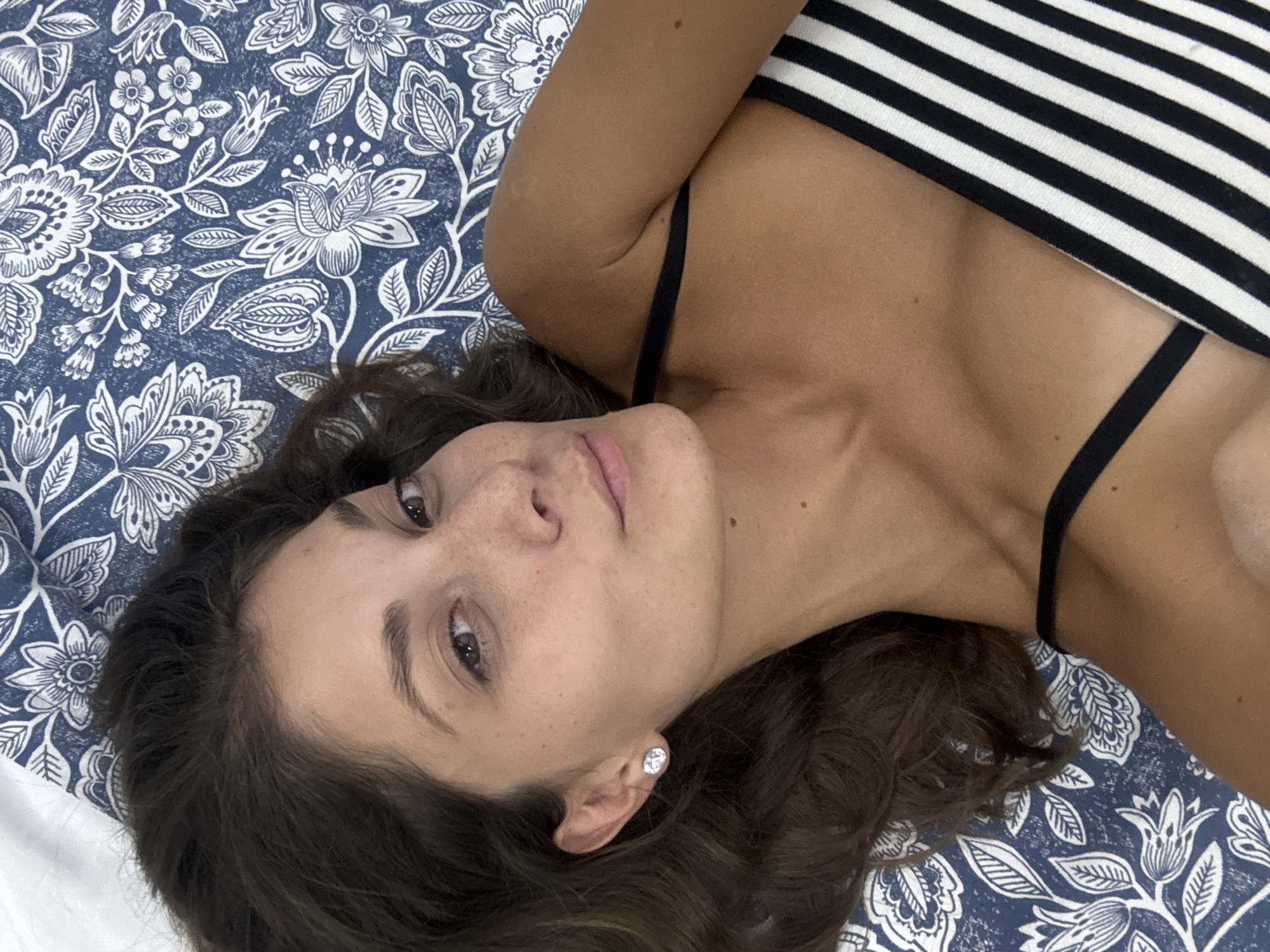 A fair-skinned woman with brown hair and freckles lies on a blue and white floral patterned surface. She wears a black and white striped top with thin black straps. Her head is tilted, with her right ear visible, adorned with a stud earring. The context does not provide information relating to Kuala Lumpur or other places.