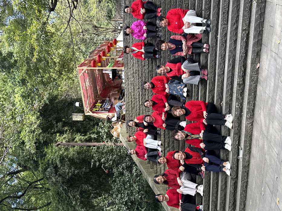 The image captures approximately 20 individuals, primarily women, situated on a broad stone staircase within a lush green environment in Yuzhong District, China. Nineteen women, predominantly appearing middle-aged to elderly, are seated on various steps of the staircase, facing forward, indicating a posed group photograph. Most are attired in red tops, ranging from sweaters to jackets, combined with pants or skirts of varying colors. One woman among this group wears a bright pink jacket.
In the upper-left portion of the image, positioned at the top of the staircase, a temporary market stall with a red and white patterned fabric canopy and a wooden frame is visible. Several red Chinese lanterns with yellow tassels hang from the front of the stall. A younger woman is seated on a chair within the stall, behind a counter, suggesting an attendant or vendor. Chinese characters are visible on a sign inside the stall.
The background consists of dense green foliage, including large trees and bamboo. A tall, slender wooden pole supporting a decorative lantern is positioned to the left of the market stall. The stone steps are textured and gray. A small, metallic, spring-like object rests on the ground in the bottom-center foreground.