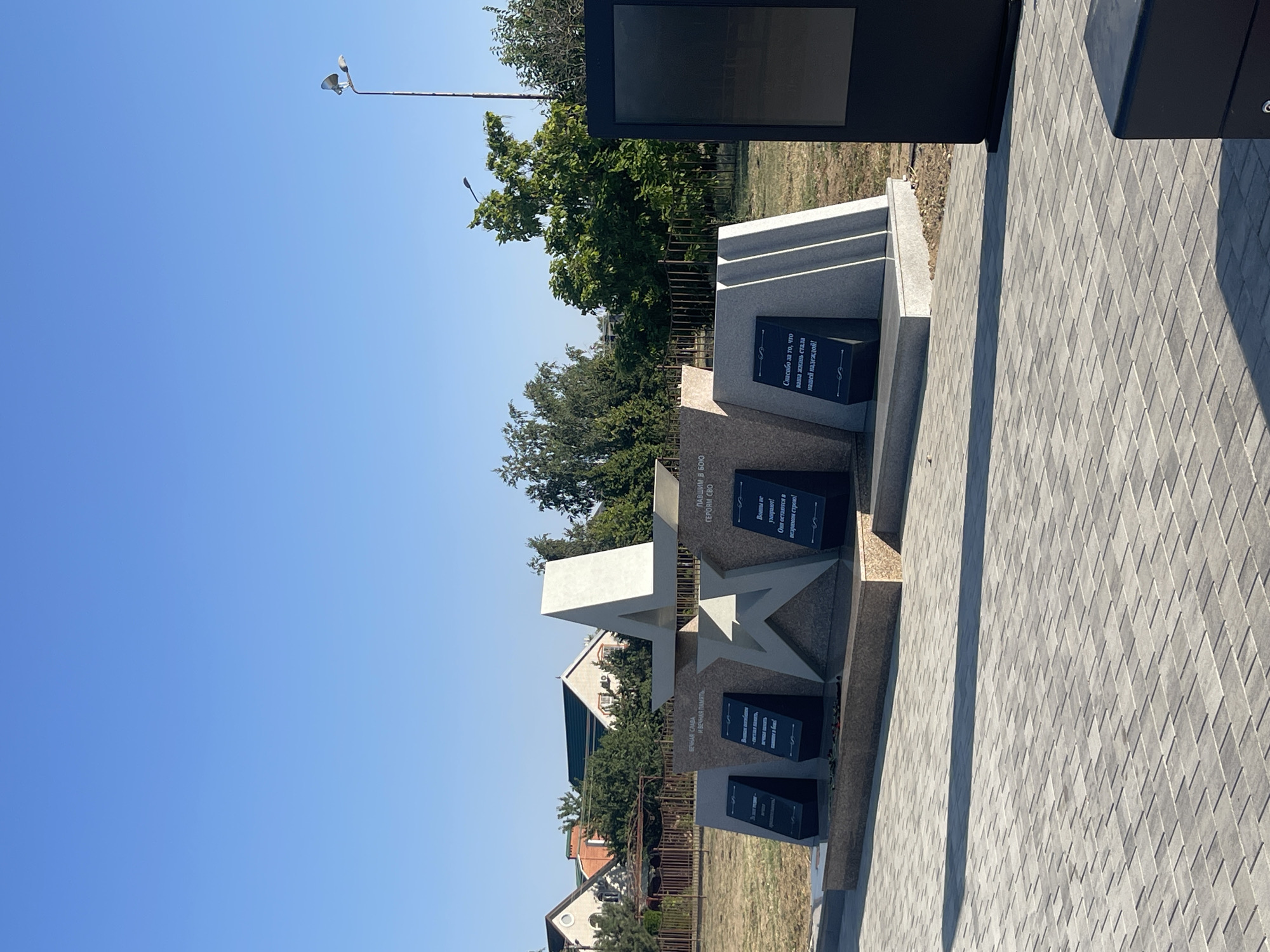 The image depicts a war memorial with star and obelisk elements, likely dedicated to soldiers. Inscribed plaques are visible on the monument, featuring text in a Cyrillic alphabet. The monument is situated in a paved outdoor area, with a fence and greenery in the background, along with residential buildings. The sky is clear and blue, indicating a sunny day. The location is identified as the City of Budyonnovsk, Russia.