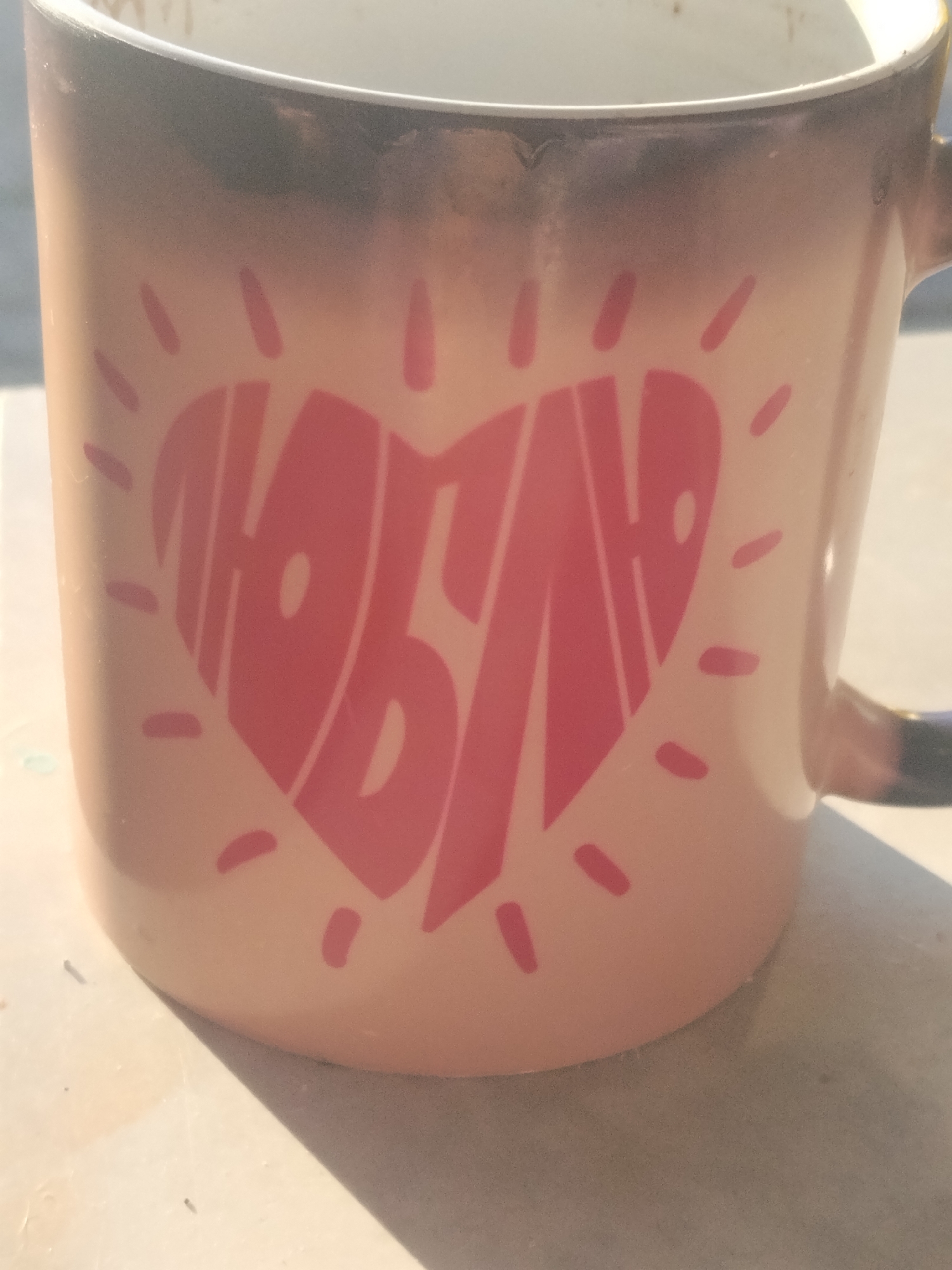 A close-up, overhead shot captures a pink mug adorned with a large, stylized heart shape containing the Cyrillic word "ЛЮБЛЮ" (Lyublyu), meaning "I love." The mug has a gradient effect at the top, transitioning from a dark, almost black color to a lighter pink. It rests on a light-colored, textured surface, possibly a table, with a distinct shadow cast across it. The background is blurred, suggesting an outdoor setting, though no specific city landmarks are visible to confirm its location in Homyel, Belarus. The lighting indicates it is daytime, with sunlight illuminating the mug.