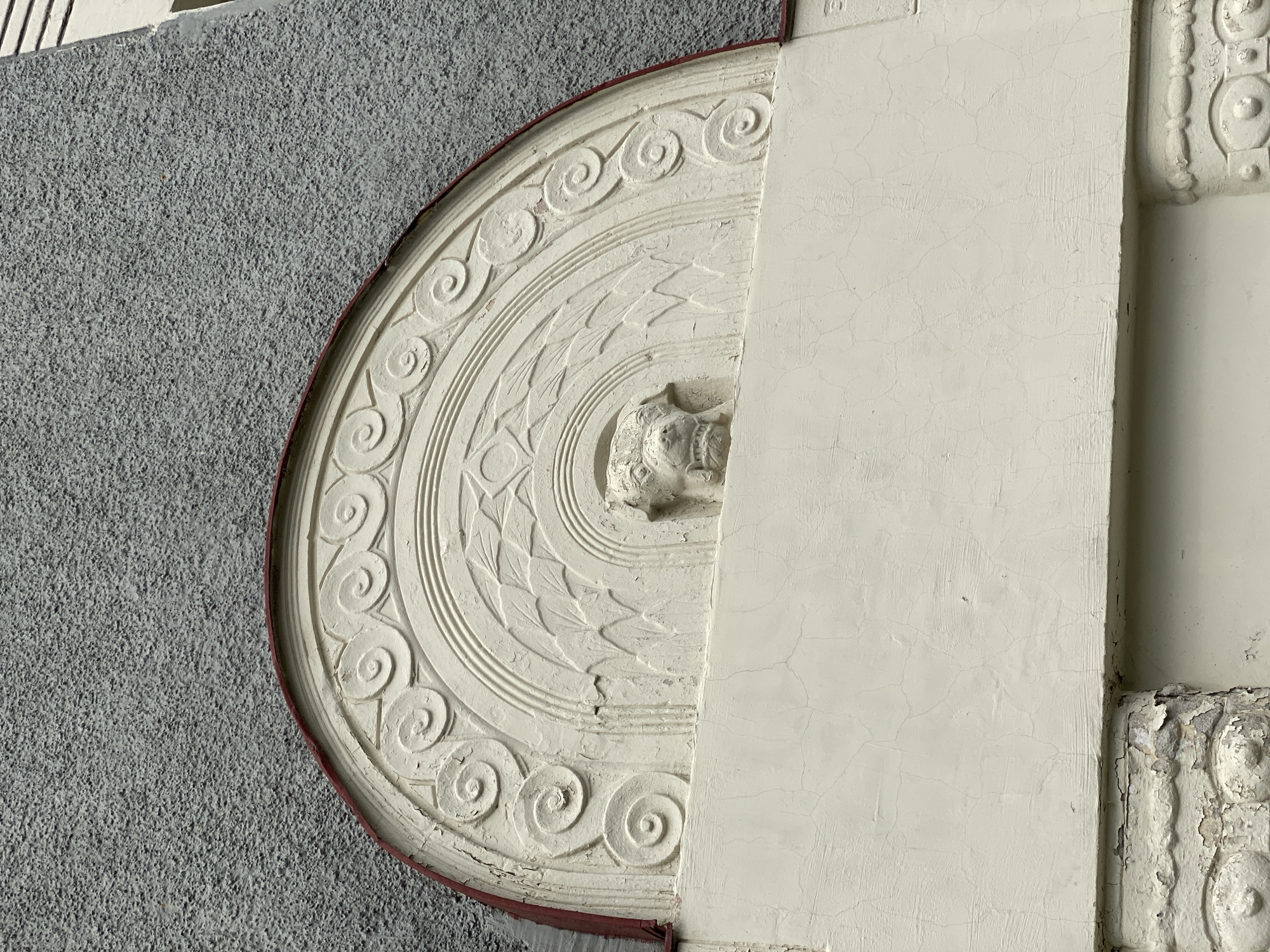 This is a close-up, eye-level, outdoor photograph of architectural ornamentation on a building in Sortavala, Russia. The subject is a carved relief in a light color, possibly stucco or plaster, set against a textured, gray exterior wall. The relief is semicircular and features a central depiction of an animal's head, possibly a lion or dog, with an open mouth and prominent teeth. Surrounding the head are concentric semi-circular bands with decorative patterns, including swirling motifs and geometric shapes resembling leaves or feathers. The relief is partially weathered, with some cracking and peeling of the surface. A thin, dark red band borders the top edge of the relief. Below the relief, a smooth, light-colored horizontal surface extends across the frame, also showing signs of cracking. The bottom of the image reveals further decorative elements with a similar worn appearance. The environment appears to be overcast, judging by the diffuse lighting. There are no people or animals present in the image.
