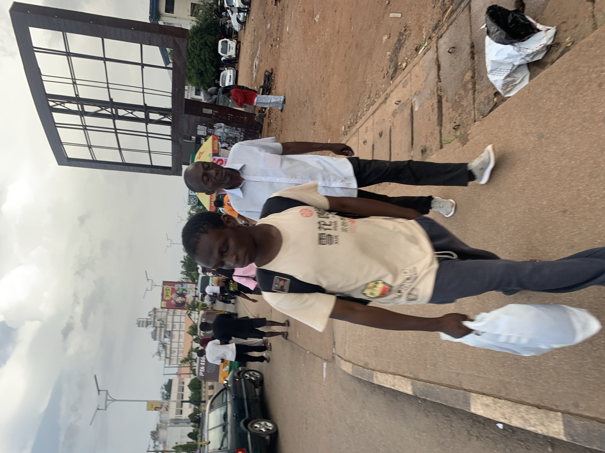 The image captures a street scene in Jos, Nigeria, under an overcast sky. In the foreground, two men are walking on a paved sidewalk. The man on the left is younger, wearing a light-colored t-shirt with graphics and dark pants, carrying a white plastic bag in his left hand. He has a backpack on his shoulders. The man on the right is older, dressed in a light blue collared shirt and dark pants. They are walking in the same direction, towards the viewer.
The background shows a street with various vehicles, including cars and what appears to be a tricycle-taxi. Several other pedestrians are visible on the street and sidewalk, some walking away from the camera. Buildings line the street, and a large, dark, metal billboard frame stands prominently in the middle ground. The ground is a mix of dirt and pavement, with some visible drainage covers. There is also litter on the side of the road, including a crumpled white and black bag. The overall impression is of a busy urban environment.