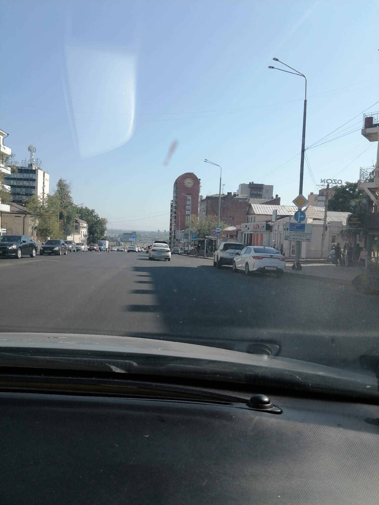 The image shows a view from inside a car looking out at a city street scene in Rostov-on-Don, Russia. The street is lined with buildings of varying architectural styles and heights, and there are several cars driving on the road. A prominent landmark is a tall, reddish-brown building with a clock tower visible on the right side of the image. There are also streetlights and traffic signs. The sky is clear and blue. The image appears to be taken on a sunny day.