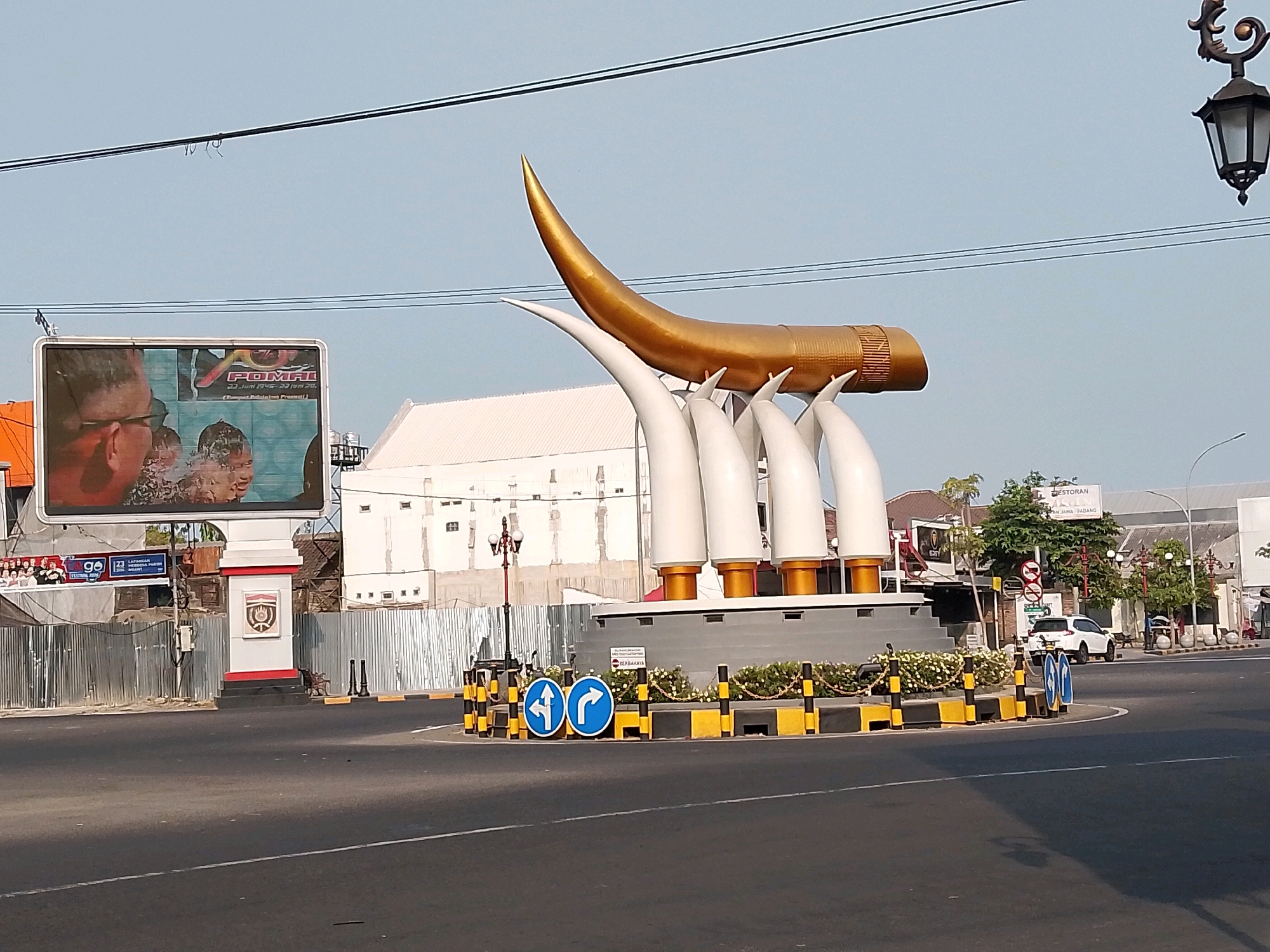 The image shows a roundabout in the city of Ngawi, Indonesia. The central feature of the roundabout is a large monument composed of several large, white, tusk-like structures supporting a golden, horn-like object. The monument is surrounded by a low wall decorated with yellow and black bollards and some small plants.
To the left of the monument, a large digital billboard displays an image, and below it, a smaller billboard with text is visible. A building with a white facade is visible behind the monument. In the foreground, there is a paved road, indicating this is a traffic intersection. Traffic signs, including directional arrows and a prohibition sign, are visible around the roundabout.
In the background, there are more buildings, trees, and a white car driving on the road. A decorative street lamp is visible in the upper right of the frame. The sky is clear and blue. The overall scene depicts a public space within a city, likely a busy intersection with a prominent landmark.