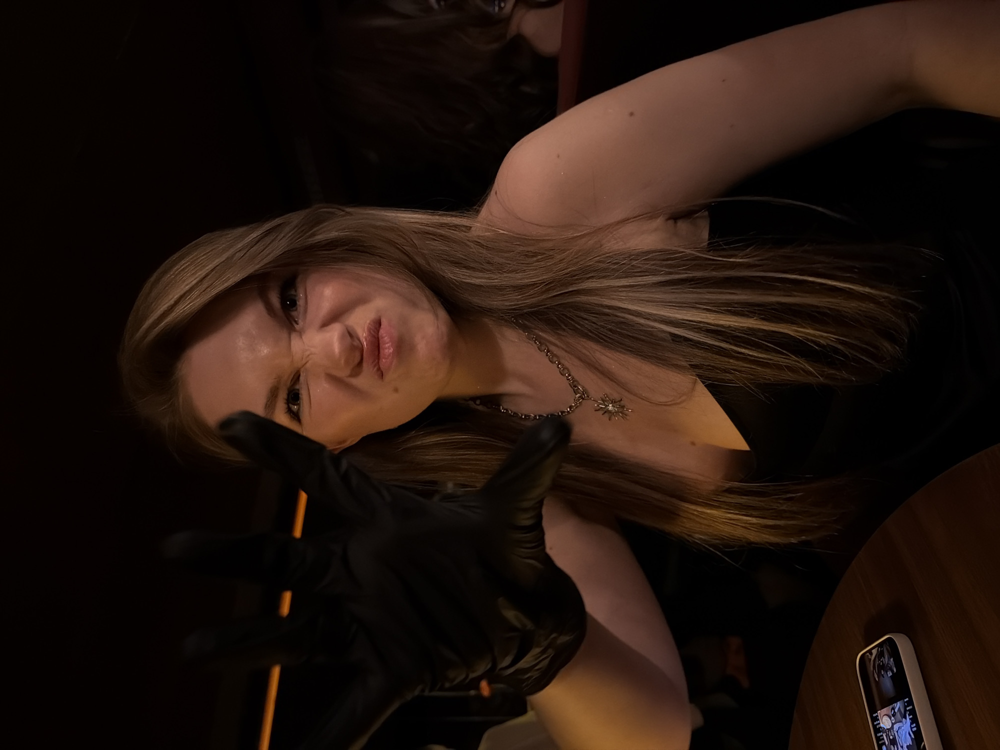 A woman with long blonde hair is posing with a black glove, making a clawing gesture towards the camera. She is wearing a dark dress and a necklace with a sun-like pendant. The background is dark, suggesting an indoor setting. There is a wooden table visible in the lower right corner with a smartphone on it. The location is likely in Belgrade, Serbia.