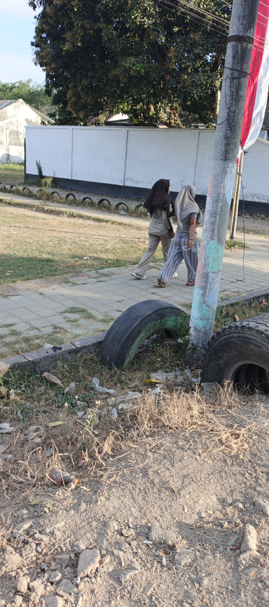 Two individuals wearing hijabs walk along a paved walkway. The person on the left wears a brown hijab and a tan outfit. The person on the right wears a lighter colored hijab and a gray t-shirt with striped pants. They are walking away from the camera. In the foreground, there are two large discarded tires, one of which is partially visible on the right side of the image. The ground around the tires is covered in dry grass and litter. Behind the walkway, there is a low white wall and a large tree with dense foliage. A light pole with some colorful paint markings and wires is visible to the right of the individuals. In the background, there are some buildings and a red and white flag attached to the pole. The scene appears to be an outdoor area, possibly a park or a public space, with signs of neglect due to the presence of discarded tires and litter. The location is Kasiyan Timur, Indonesia.