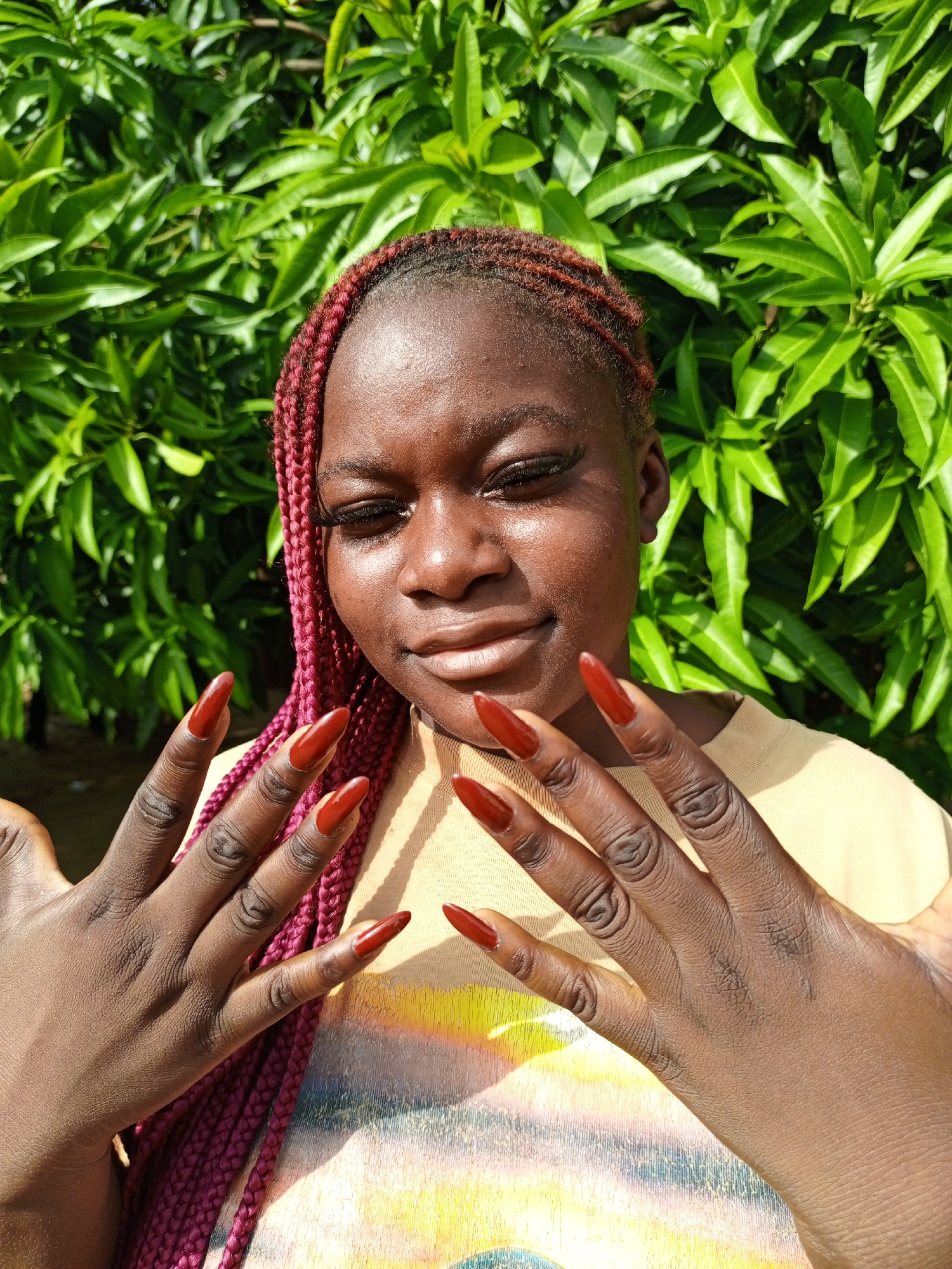 A young woman with dark skin is pictured outdoors in Keffi, Nigeria, posing with a subtle smile. Her hair is styled in reddish-brown or burgundy braided cornrows. She wears a light-colored t-shirt with a multi-colored graphic design visible on the chest.
The woman holds both hands up near her chest, prominently displaying long, pointed nails painted in a glossy, deep red color. Her fingers show distinct skin texture and creases. Her face is brightened by strong, direct sunlight, which also creates highlights on her hair and nails. Her eyelashes appear dark and full.
The background is composed entirely of lush, vibrant green foliage, suggesting a natural, outdoor setting, possibly a garden or a bush. The bright, direct light indicates it is daytime with sunny weather. The overall mood is calm and confident, with the woman seemingly showcasing her manicure. There is no visible text in the image.