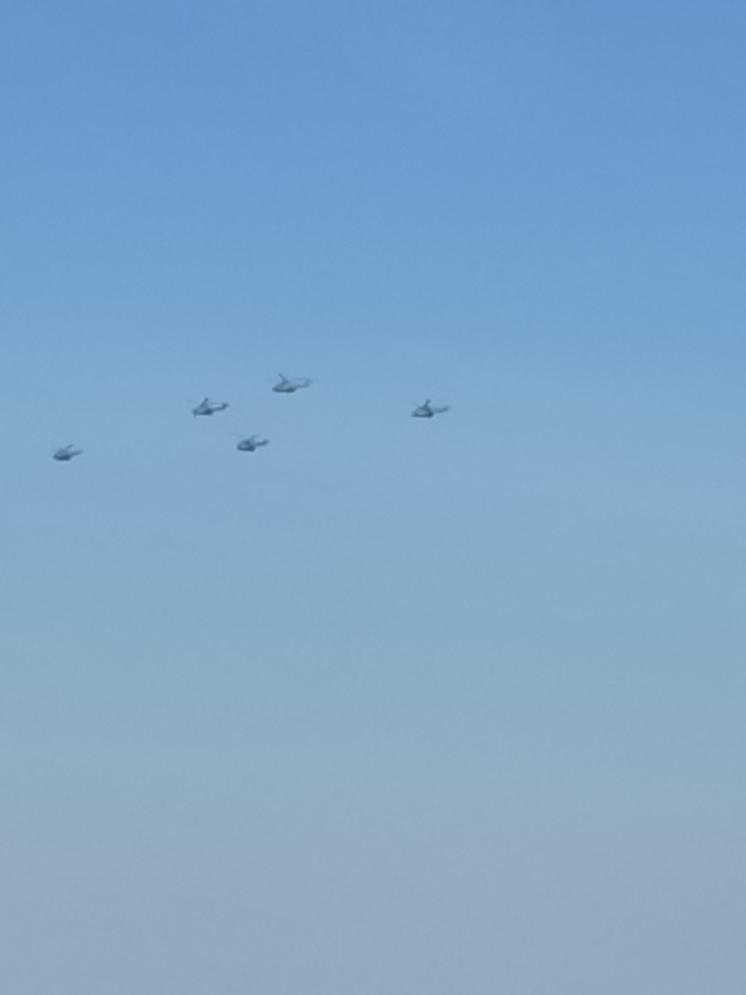 In the bright, clear sky above Crețuleasca, Romania, a formation of five helicopters is captured mid-flight. The helicopters are spaced apart in a loose V-shape, suggesting a planned aerial maneuver. The sky is a uniform light blue, indicating fair weather conditions. The helicopters themselves are small specks against the vast expanse of the sky, but their distinct shapes, with rotors and fuselages, are discernible. Their position suggests they are flying at a significant altitude. The scene is devoid of any other objects or people, focusing solely on the aircraft against the natural backdrop of the sky.