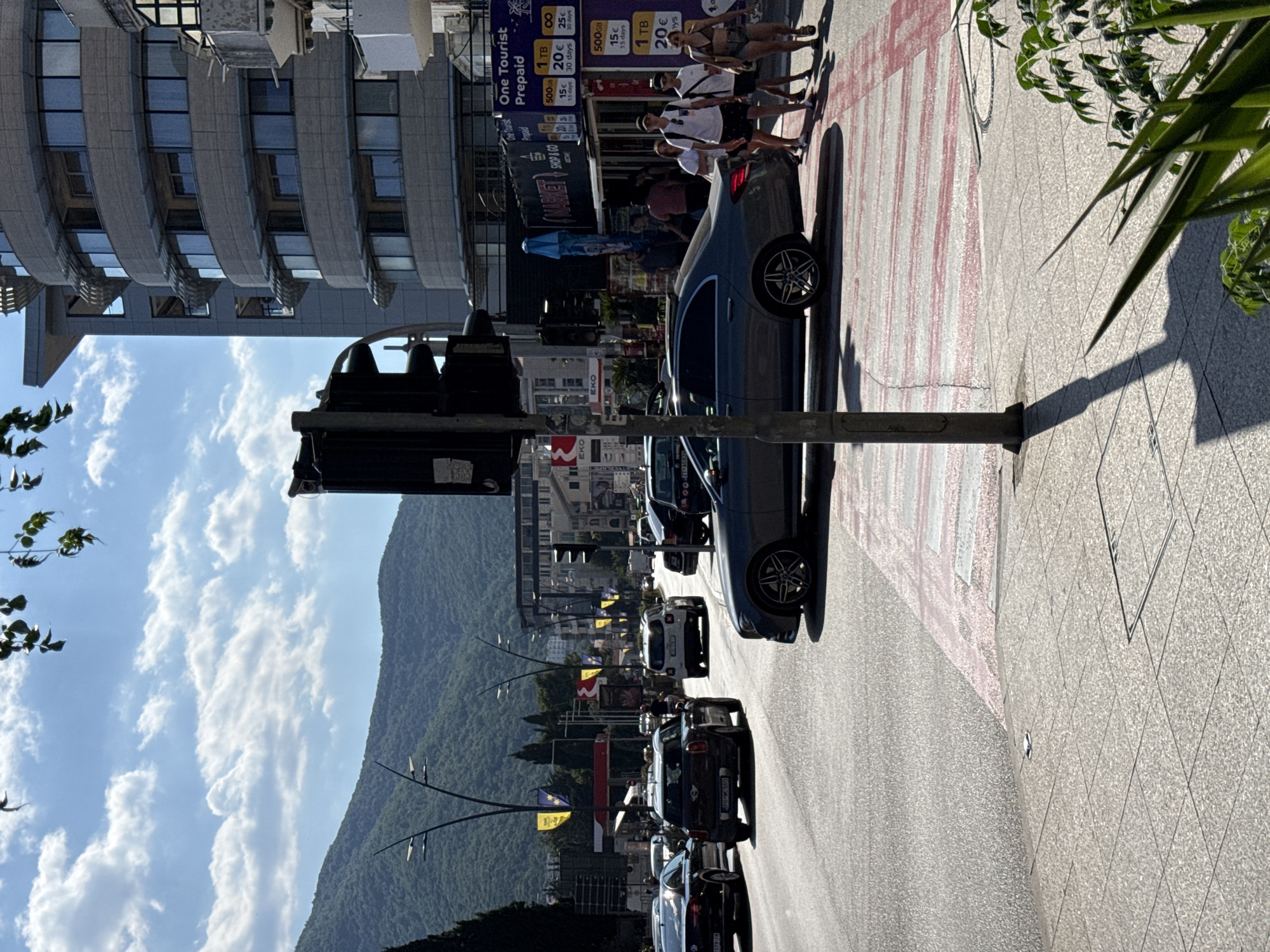 The image depicts a street scene in Budva, Montenegro, likely during the daytime given the bright sunlight and shadows. Several cars are parked or driving along the road, including a dark-colored sedan in the foreground. A group of people, appearing to be tourists, are walking on the sidewalk, dressed in casual summer attire. They seem relaxed and are interacting with each other.
Prominent signs are visible along the street, including "EKO" and "MARKET SHOP & GO". One sign advertises "One Tourist Prepaid" mobile data plans, with options for 500GB and 1TB at various prices and durations. These details suggest a commercial area catering to tourists.
The background features a modern building with numerous windows and a steep, tree-covered mountain rising behind the town. The sky is partly cloudy with fluffy white clouds scattered across a clear blue expanse. The overall atmosphere is bright and sunny, indicating pleasant weather suitable for outdoor activities. The perspective is from street level, looking down a road that curves slightly. The presence of traffic lights and pedestrian crossings suggests this is a busy urban thoroughfare.