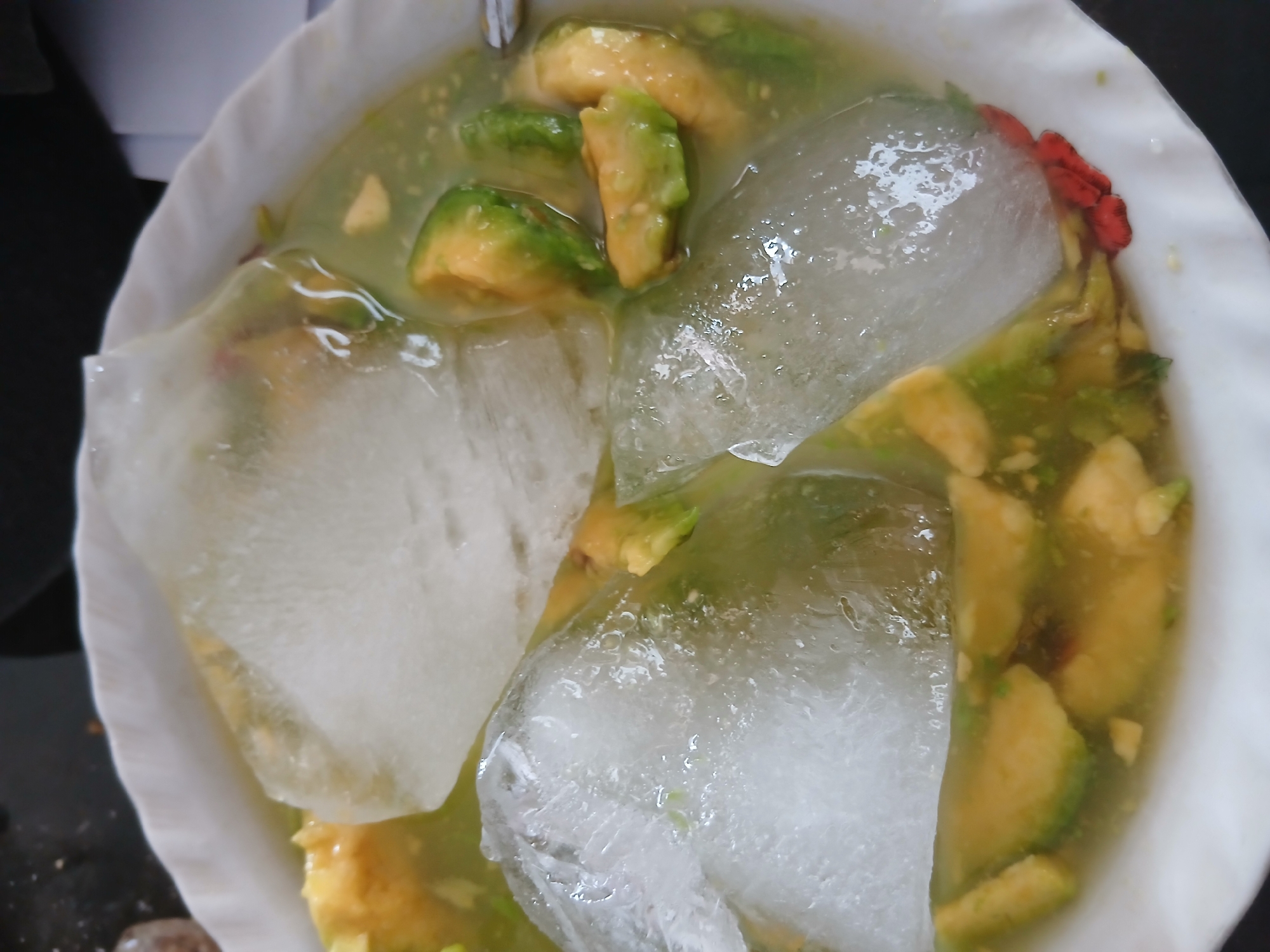 In a white bowl, sliced avocado is submerged in a pale green liquid, possibly a soup or dessert. Large pieces of ice float on top, some with rough edges and others appearing smoother and more melted. The avocado slices show a mix of green skin and yellowish-green flesh. The liquid appears somewhat opaque, with small specks visible within it. The bowl has a slightly ruffled edge. The background is dark and out of focus, suggesting the image was taken indoors. No people or specific environmental details related to Tenggarang, Indonesia, are discernible in this close-up shot of the food.