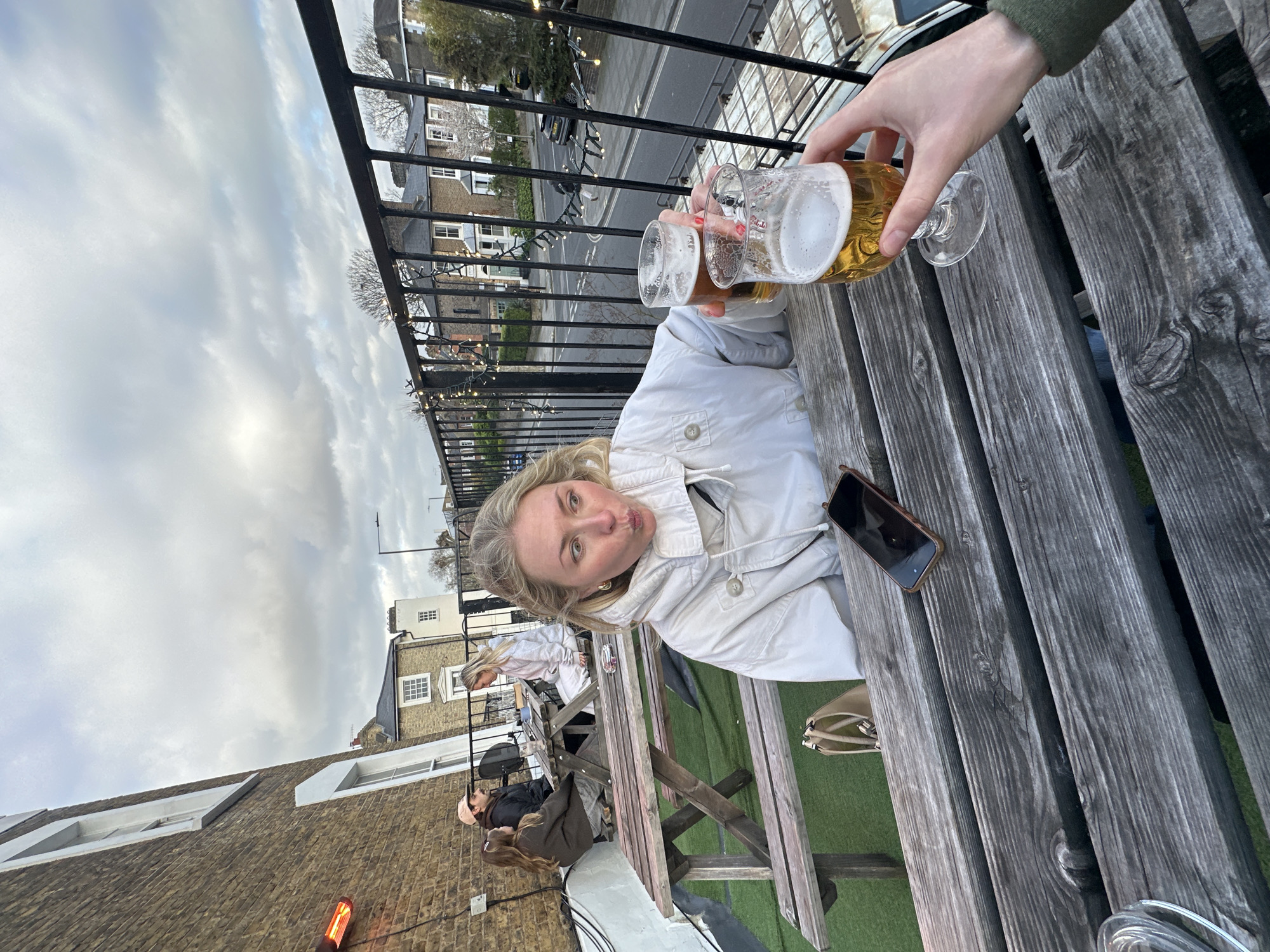 The image captures a social scene likely in London, United Kingdom. A woman with blonde hair, wearing a light-colored jacket, poses for the camera with her lips puckered, appearing playful. She is seated at a wooden picnic table outdoors.
On the table, there is a mobile phone placed next to her. A person's hand extends into the frame holding a pint of beer towards the woman, possibly for a toast. Another pint of beer can be seen being held by someone out of frame.
Other people are seated at the same table but are further away from the camera and less prominent. In the background, there is a street scene with buildings typical of London architecture. A black iron railing separates the outdoor seating area from the street. The sky is overcast with grey clouds.