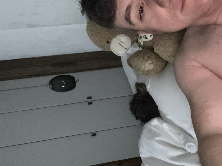 The image displays an indoor bedroom scene in low light. A male subject is positioned in the foreground on the right, shirtless, with his right eye and partial face visible, directed towards the camera. To his left, the back of a second individual's head, featuring dark hair gathered in a bun, is visible resting on a white pillow under white bedding, indicating they are asleep. A large, light brown stuffed animal with a lighter snout and a dark-and-white patterned ribbon around its neck is situated between the two individuals. In the background, a white multi-panel wardrobe with dark handles is visible. A dark wooden vertical structural element is present to the right of the wardrobe. A dark, elongated, oval-shaped pendant light fixture is suspended from the ceiling in the upper-middle background. The specific location within Kastelruth - Castelrotto, Italy, cannot be determined from this image.