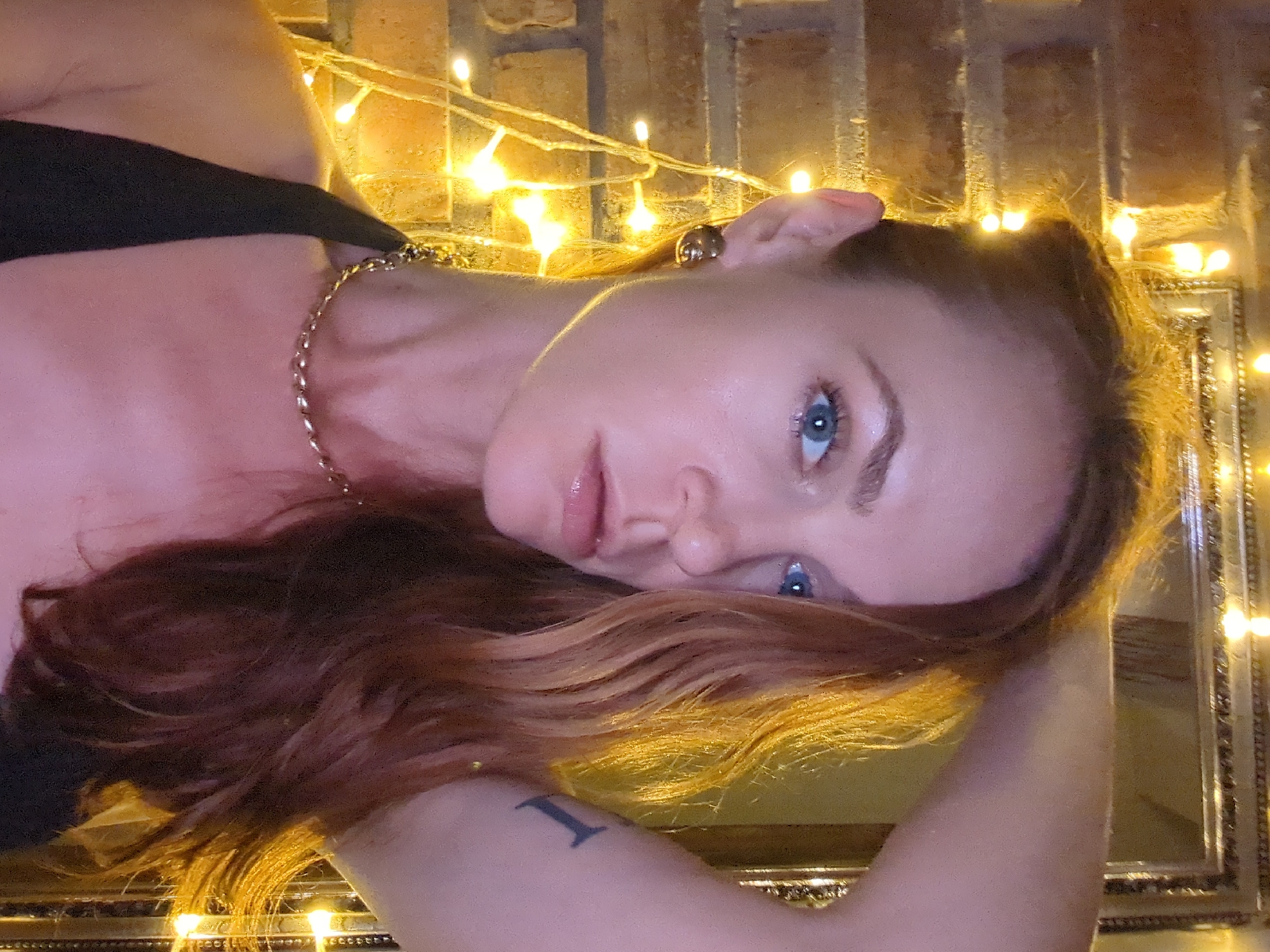 The image, rotated 90 degrees clockwise, portrays a woman with fair skin, blue eyes, and long, wavy reddish-brown hair with golden highlights. She looks directly at the camera with a neutral expression. She is wearing a dark top and accessorized with a gold-toned chunky chain necklace and a gold-toned earring visible on her right ear. A dark tattoo, appearing to be the Roman numeral 'I' or a capital 'I', is visible on her left upper arm.
The background features a brick wall, softly illuminated by numerous warm, glowing fairy lights, some of which are blurred to create a bokeh effect. These lights serve as the primary illumination, suggesting an evening or nighttime setting. A decorative gold-framed mirror is visible on the right and bottom edges of the frame, reflecting the warm glow of the fairy lights. The woman's hair also catches the golden light, adding to the intimate and warmly lit atmosphere.