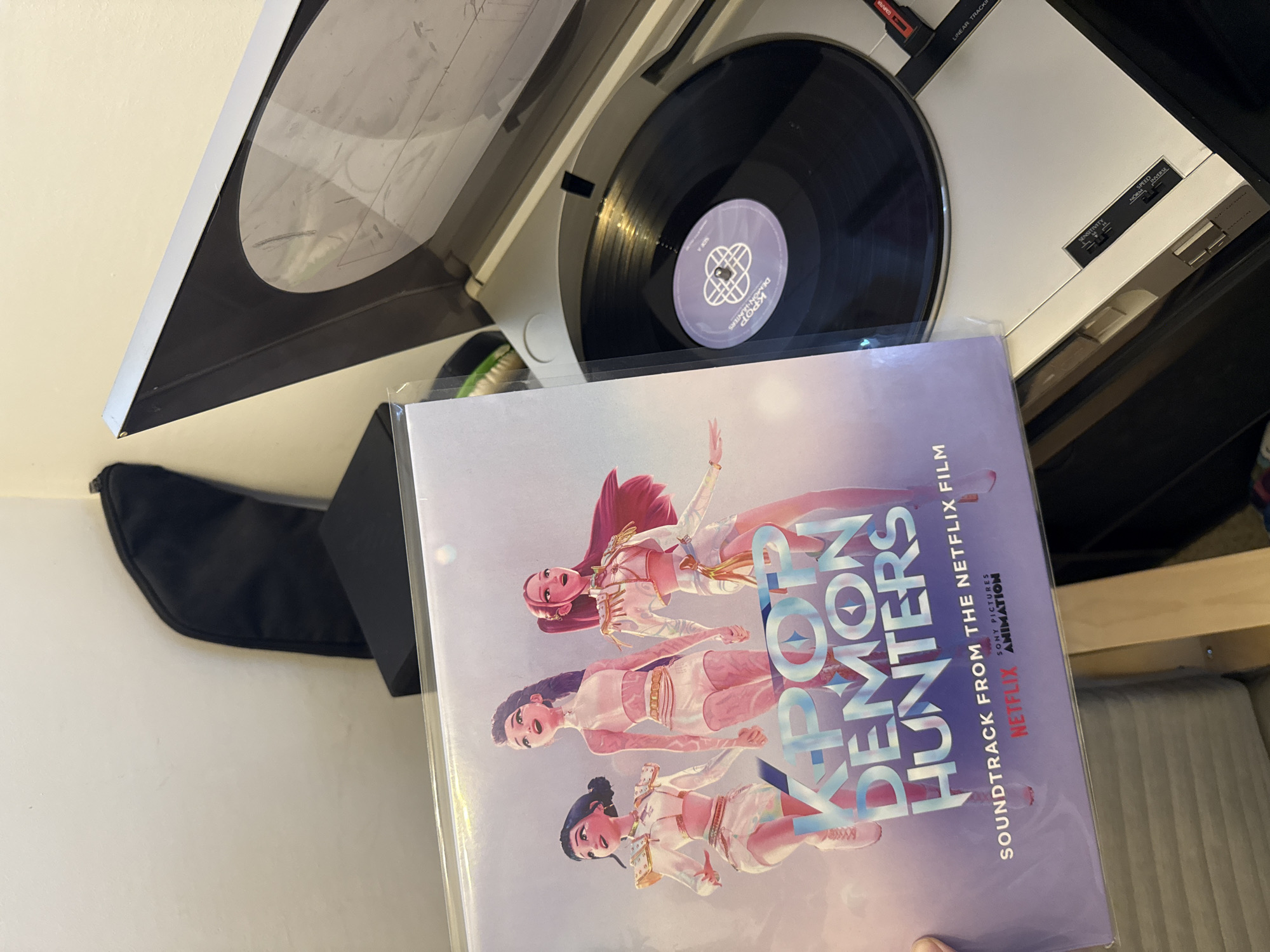 An overhead, angled shot captures a vinyl record setup with an album cover in the foreground. A light-colored record player with a black platter and a clear, partially open dust cover occupies the upper half of the image. A black vinyl record with a purple label featuring "K-POP DEMON HUNTERS" and "SIDE A" is visible on the turntable. The record player's controls are partially visible, including text like "SENSITIVITY", "NOR", "SPEED", and "INVERSE", along with "LINEAR TRACKING".
In the foreground, a hand holds a glossy, light purple vinyl album cover for "K-POP DEMON HUNTERS", which is "SOUNDTRACK FROM THE NETFLIX FILM". The cover art depicts three animated female characters in dynamic poses, dressed in stylized light-colored outfits. Additional text on the album cover includes "NETFLIX", "SONY PICTURES", and "ANIMATION".
The background features a plain light-colored wall on the left, with a black soft case or bag resting against it. On the right, a wooden frame and upholstered furniture piece are partially visible. The scene suggests an indoor setting, likely a home, with someone preparing to play or having just played a vinyl record. This activity is taking place in Petah Tikva, Israel.