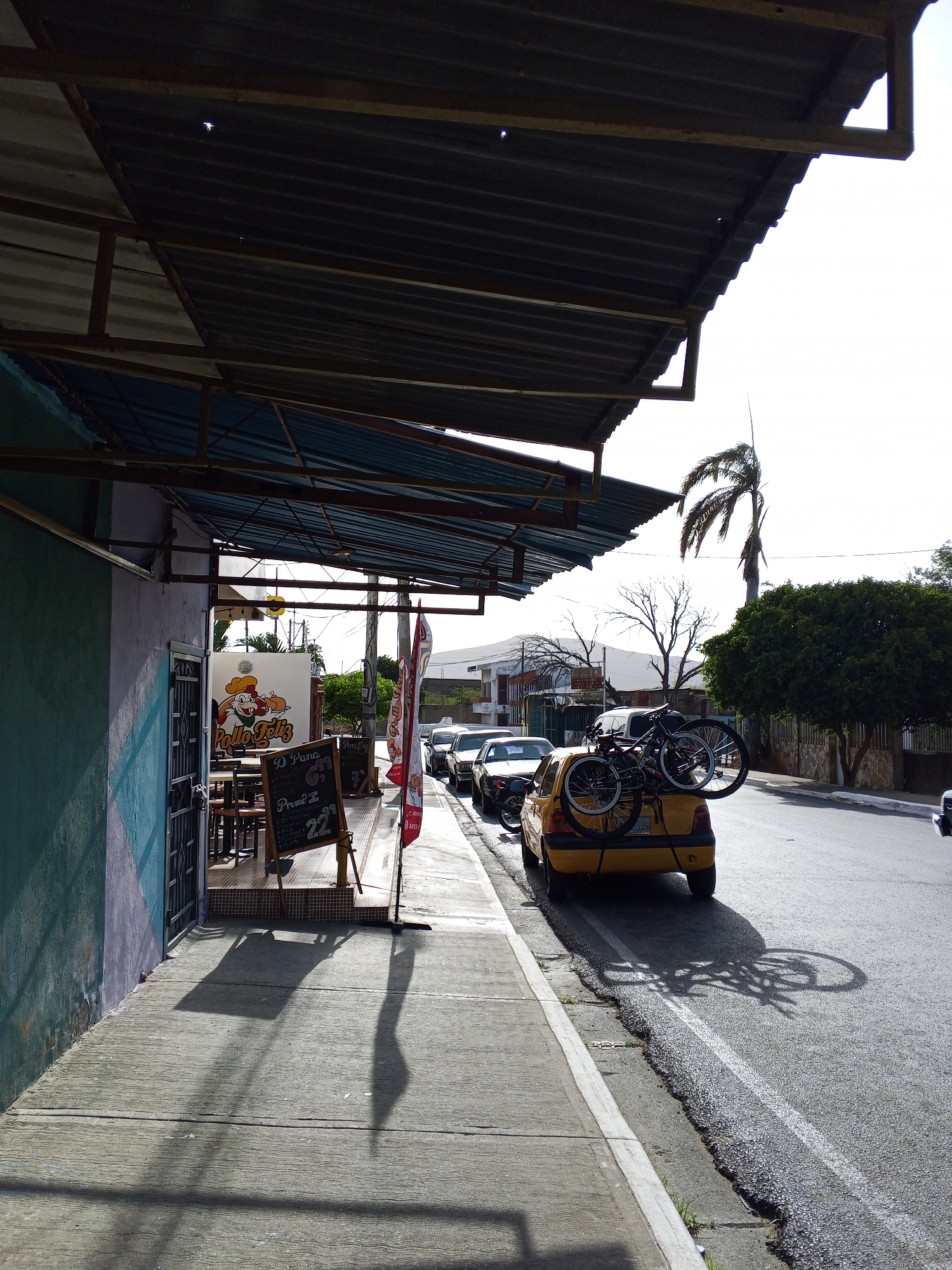 The image shows a street scene in an urban setting in an unidentified city. A yellow car is parked on the right side of the street with two bicycles secured on its rear. There are other cars parked in a line along the curb in the background. A sidewalk runs along the left side of the street, in front of a building with a covered outdoor seating area. The building appears to be a restaurant, with a sign that reads "Pollo Feliz" and a menu board displaying prices. Shadows from the awnings and signs are cast onto the sidewalk. A palm tree and other trees are visible in the background.