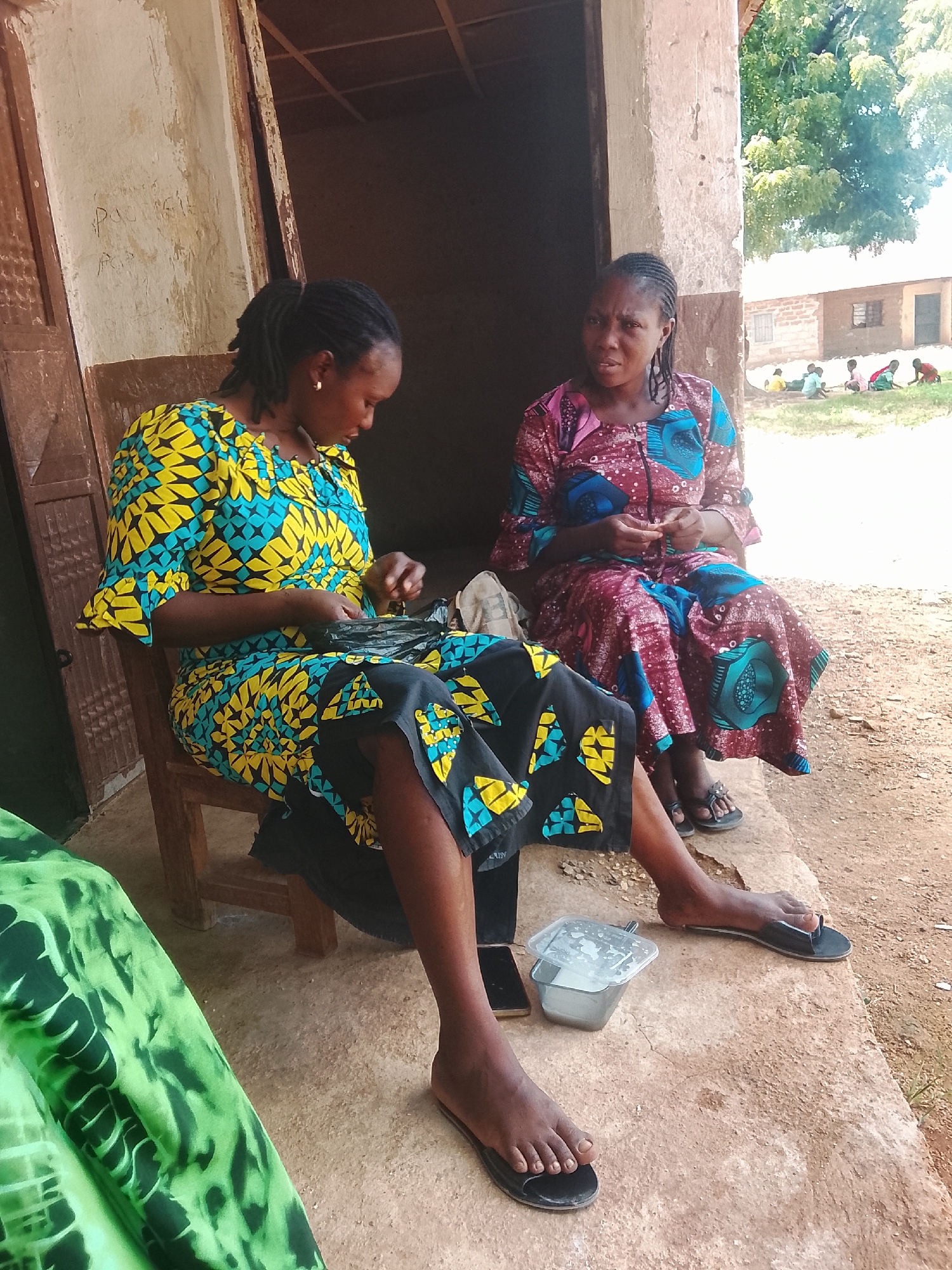 Two women are seated outside a building in Akwanga, Nigeria. The woman on the left wears a yellow and blue patterned dress and is looking down, appearing to be working with a black plastic bag. She is seated on a wooden chair. The woman on the right wears a maroon and blue patterned dress and is looking towards the viewer with a neutral expression. She is seated on a concrete ledge. In front of the woman on the right is a small, clear plastic container with a lid. A smartphone lies on the ground between the two women. The building behind them has a rough, plastered exterior with a wooden door frame. Further in the background, several other people, including children, are visible, some seated on the ground near another building. The ground is made of dirt and loose gravel, and trees and foliage are visible in the distance.