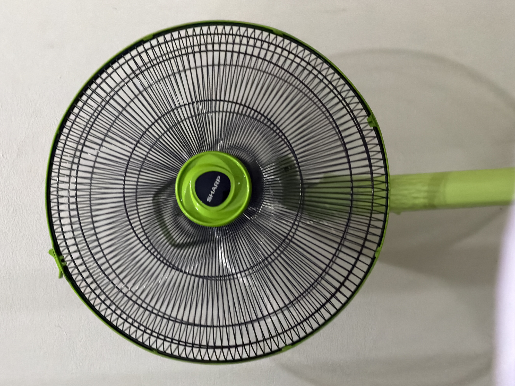 A close-up shot shows a black wire mesh fan guard with a bright green rim and center. The center has a circular black emblem with the word "SHARP" in white. The fan is mounted on a light green pole that extends downwards out of the frame. The background is a textured, off-white wall, with a shadow cast by the fan. The image is captured in Baan Mae Nam, Thailand, suggesting an indoor setting, possibly a home or business.