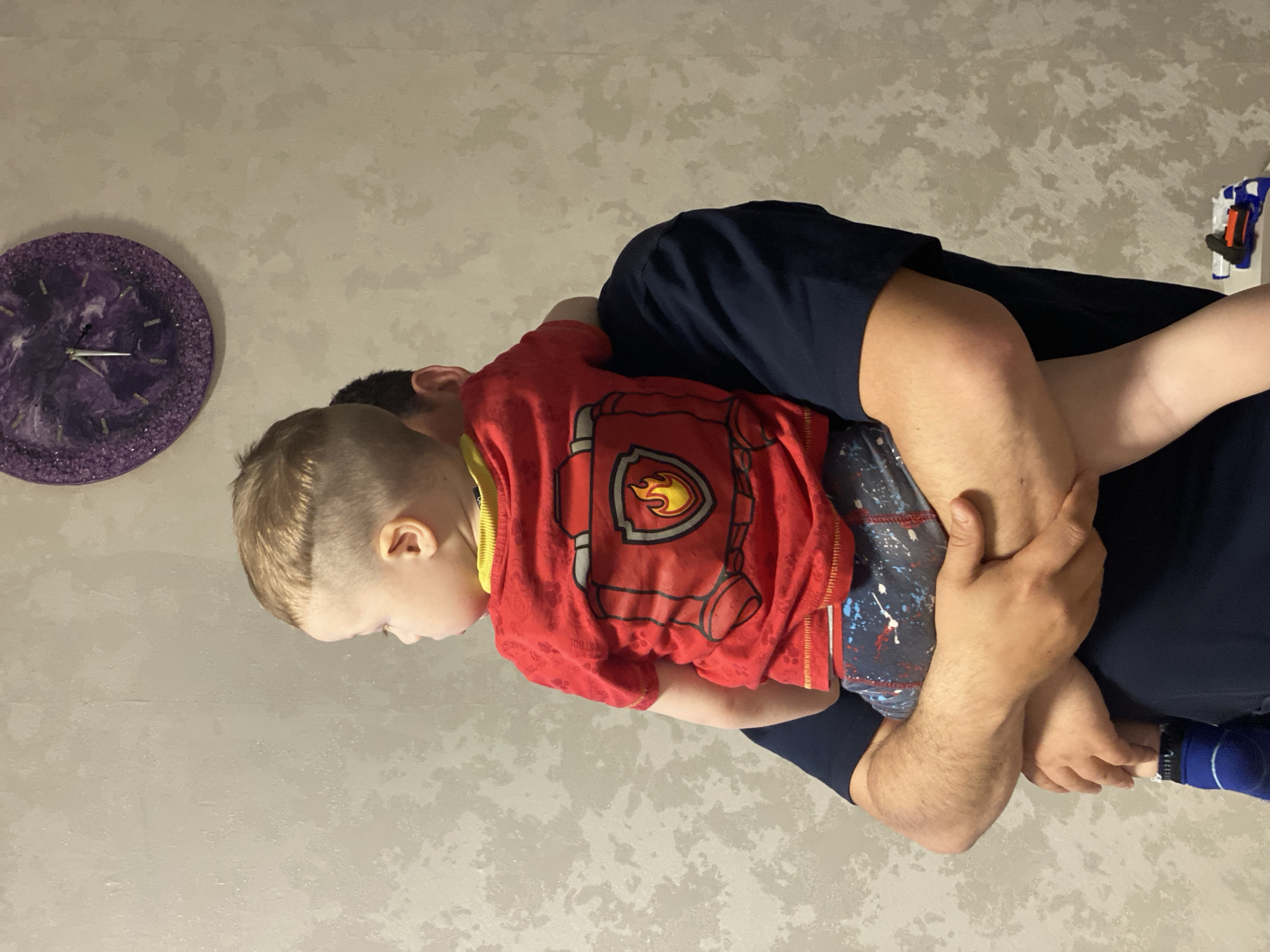 A man holds a young child in an indoor setting in Sochi, Russia. The image captures a casual moment with both individuals positioned centrally.
The man, with dark hair and wearing a navy blue t-shirt, holds the child securely in his arms. His right arm is visible, supporting the child, while his left arm and hand are also seen, providing additional support.
The child is young, with light blonde hair, and is dressed in a red t-shirt featuring a prominent Paw Patrol character shield design, likely "Marshall" the firefighter dog. The t-shirt has a yellow collar. The child is also wearing blue shorts with a white and red splatter pattern, and a blue sock on one foot. The child's head is turned, looking downwards or to the side, away from the camera.
The background consists of a light-colored wall with a subtle, mottled texture, suggesting a painted or plastered surface. On the left side of the frame, a round, purple wall clock with sparkling or textured edges and simple white hour markers is visible. The clock has silver hands. On the right side of the frame, partially visible, is a blue and orange toy gun, possibly a Nerf blaster, mounted or hanging on the wall. The scene appears to be a typical family moment, likely occurring during the day or evening indoors. No specific emotions are distinctly conveyed, giving the impression of a routine interaction between parent and child.