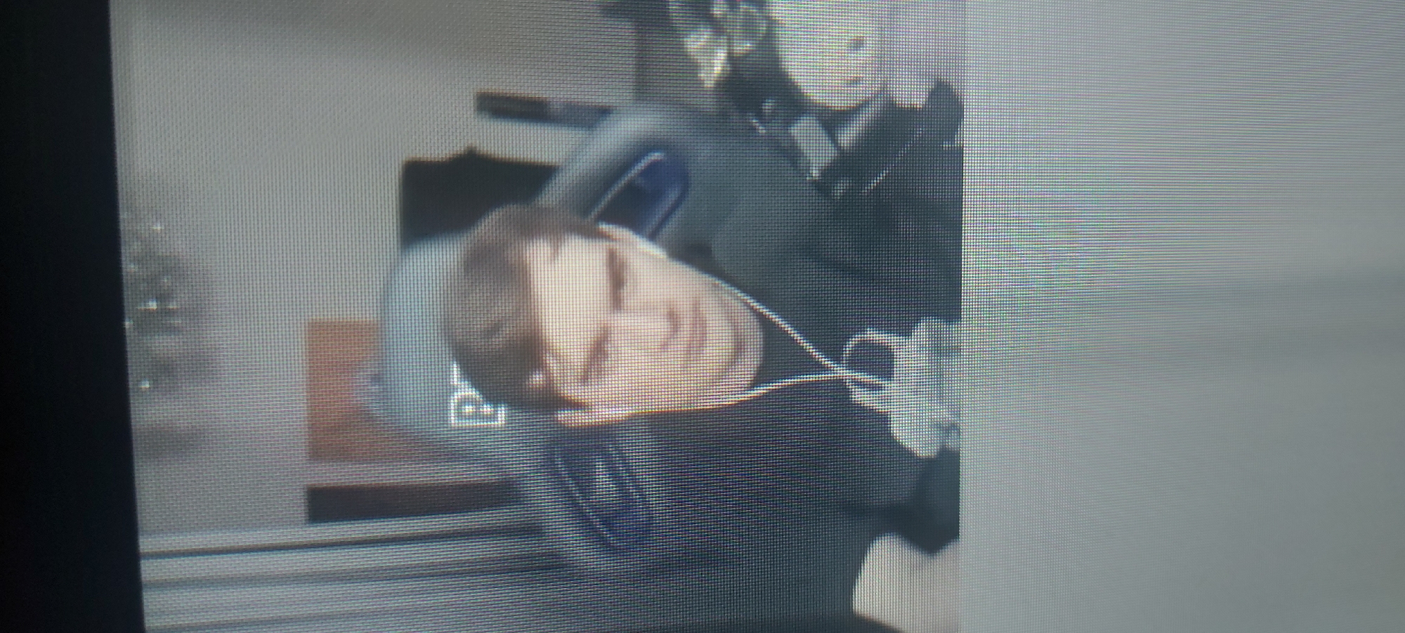 A person with short brown hair is lying on a light-colored pillow, possibly resting or sleeping. The person is wearing a dark-colored top. A second person, wearing a dark-colored outfit, is standing behind them, partly obscured. An object, possibly a headset, is visible near the first person's ear. The background is indistinct. The photograph is grainy. There is no information in the image to indicate the location of Tashkent, Uzbekistan.