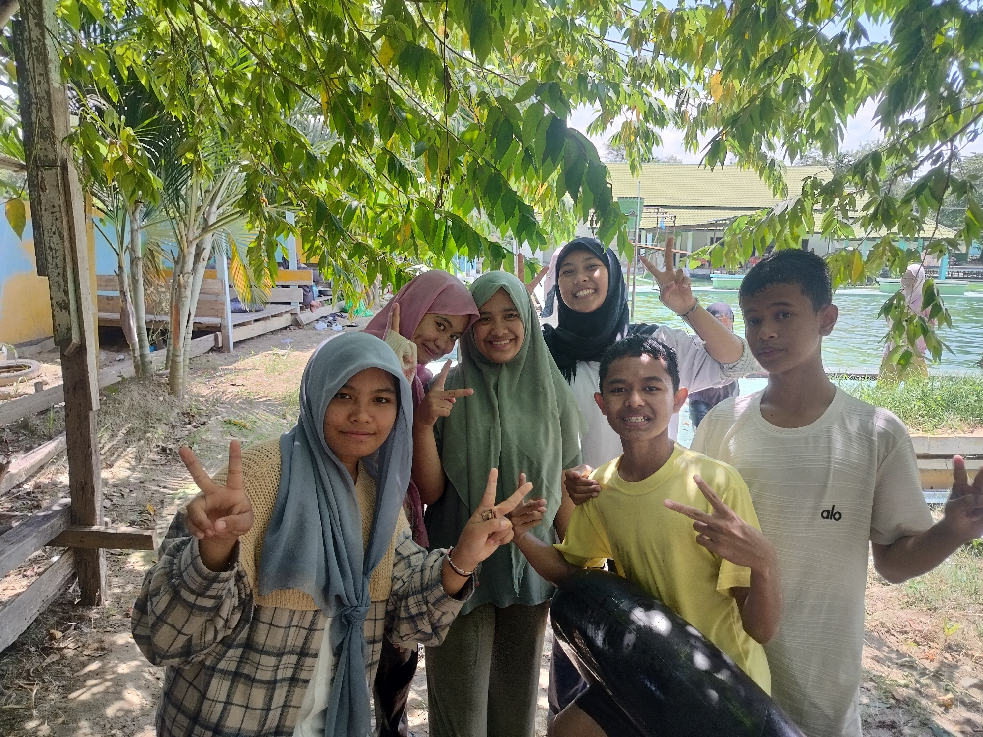 Six individuals, five of whom are teenagers, are posing for a photograph outdoors, possibly in the city of Tegarmiko, Indonesia. They are standing near a body of water, with lush green trees overhead and buildings in the background. The group consists of three young women wearing hijabs in various colors, and three young men. Two of the young women are smiling and making peace signs with their fingers. A third young woman is also smiling, and another individual is making a peace sign. One of the young men is wearing a yellow t-shirt, and the other is wearing a white t-shirt with a logo. The overall atmosphere appears casual and friendly.