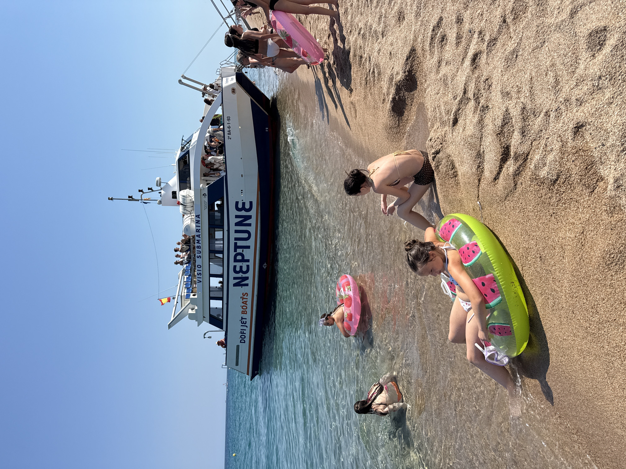 The image depicts a sunny day at a beach in Blanes, Spain. The foreground shows several young people in swimwear, some wading in the shallow, clear water and others on the sandy shore. One girl, wearing a bikini and a snorkel mask, is holding a large, inflatable watermelon-shaped raft and a snorkel set. She is sitting on the sand, with her legs in the water. Another girl, also in a bikini, is kneeling in the water, playing with an inflatable ring with strawberries on it. Further back, a tour boat named "NEPTUNE" is docked close to the shore. The boat is white with a blue stripe and appears to be a sightseeing vessel, with "VISIO SUBMARINA" and "DOFI JET BOATS" visible on its side. Several people are visible on the deck of the boat, looking out towards the water or the shore. The sky is a clear, bright blue, indicating good weather. The sunlight casts distinct shadows on the sand, suggesting it is midday. The overall atmosphere is one of a relaxed, enjoyable summer day at the beach. Visible text includes "NEPTUNE", "VISIO SUBMARINA", "DOFI JET BOATS", and a registration number "2ª BAL-6-1-93" on the boat, as well as a Spanish flag.
