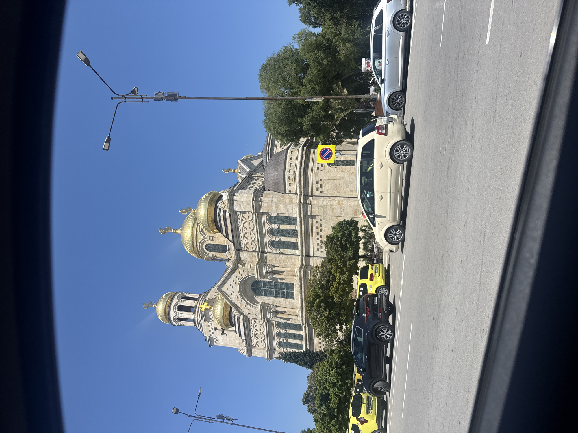In the city of Varna, Bulgaria, a view of the Dormition of the Mother of God Cathedral is captured from a street level perspective. The large, ornate cathedral, a prominent landmark in Varna, features multiple golden domes and intricate architectural details. The sky above is a clear, bright blue, indicating a sunny day. In the foreground, a street is visible with several vehicles, including yellow taxis and other cars, suggesting a bustling urban environment. The image appears to be taken from inside another vehicle, as indicated by the dark foreground which resembles the interior of a car. The scene is calm and static, with no apparent actions or interactions among the people or objects. The environment is an urban setting with a mix of historical architecture and modern transportation.