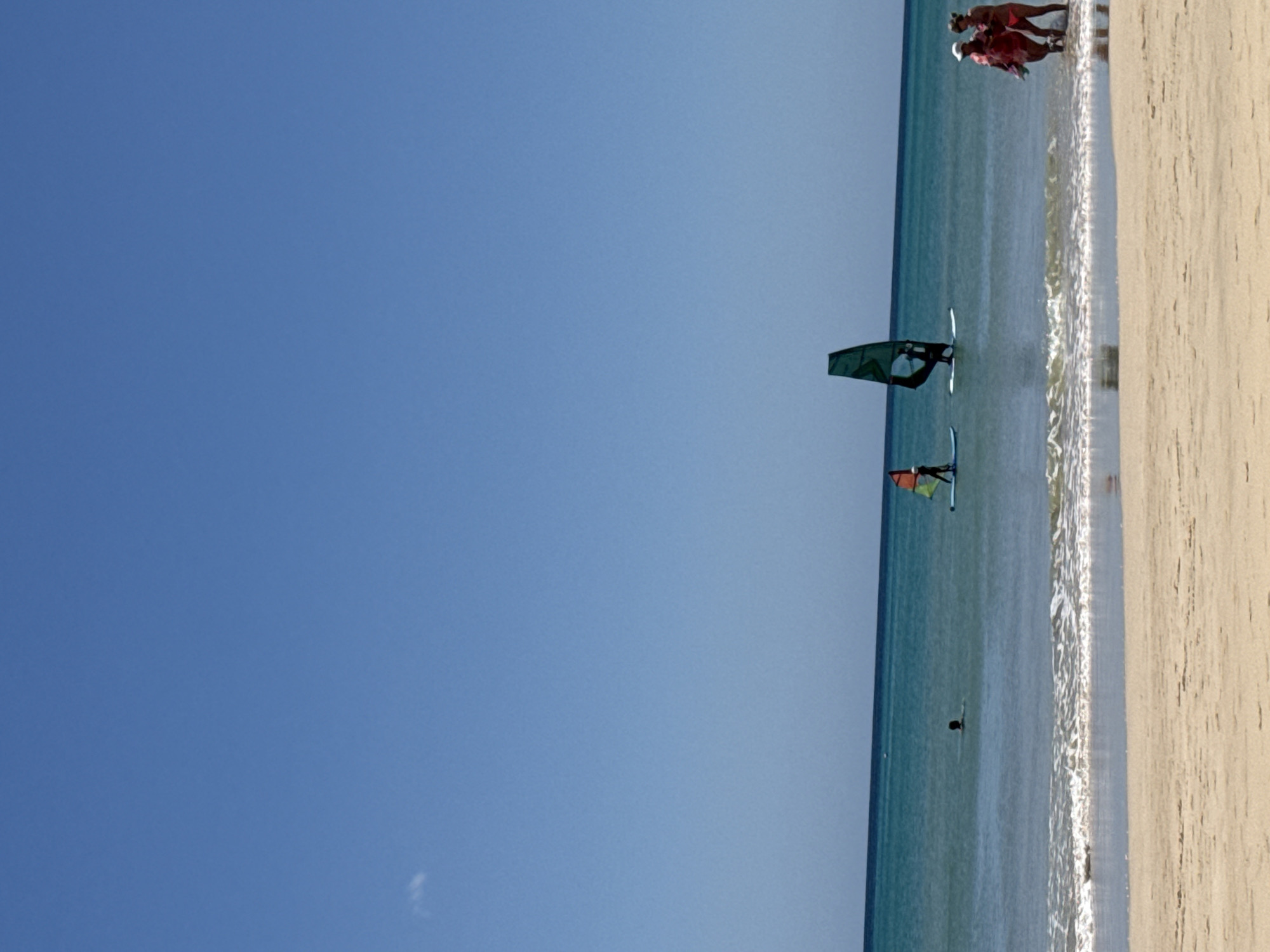 The image depicts a beach scene in Pájara, Spain. The sky is clear blue, contrasting with the tan sand of the beach on the right side of the image. In the ocean, there are two windsurfers with colorful sails. In the distance, close to the beach, a group of people are standing near the water. The water itself is a light blue, with visible waves approaching the shore. A solitary figure is seen floating further out in the water.