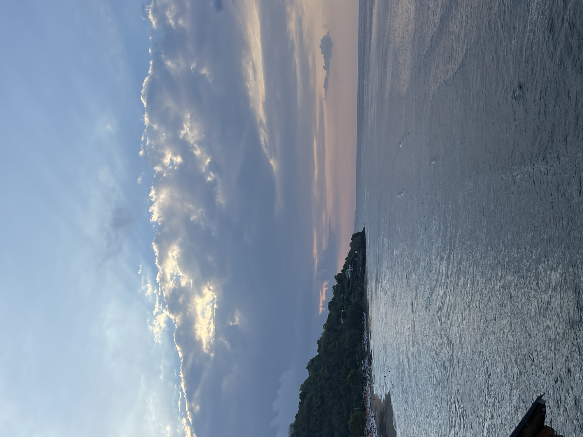 The image depicts a panoramic view of a coastline, likely in Abang, Indonesia, during what appears to be late afternoon or early evening. The sky is filled with dramatic clouds, some of which are illuminated by sunlight, creating a dynamic interplay of light and shadow. Streaks of light, known as crepuscular rays, pierce through the clouds, adding to the ethereal atmosphere.
The sea is calm, with gentle waves rolling towards the shore. The water exhibits a serene, bluish-gray hue, reflecting the colors of the sky. Along the coastline, a sandy beach is visible, lined with numerous small boats. These boats, likely traditional fishing vessels, are pulled ashore, suggesting a local community engaged in maritime activities. Some people can be seen on the beach, though their figures are small and indistinct due to the distance.
A lush, green hillside rises steeply from the beach, covered in dense vegetation. Small buildings, possibly homes or resorts, are nestled among the trees on the hillside, indicating human habitation and development. The overall scene conveys a sense of tranquility and natural beauty, characteristic of a tropical coastal region. The limited visibility of people and the focus on natural elements suggest a peaceful, undisturbed environment.
