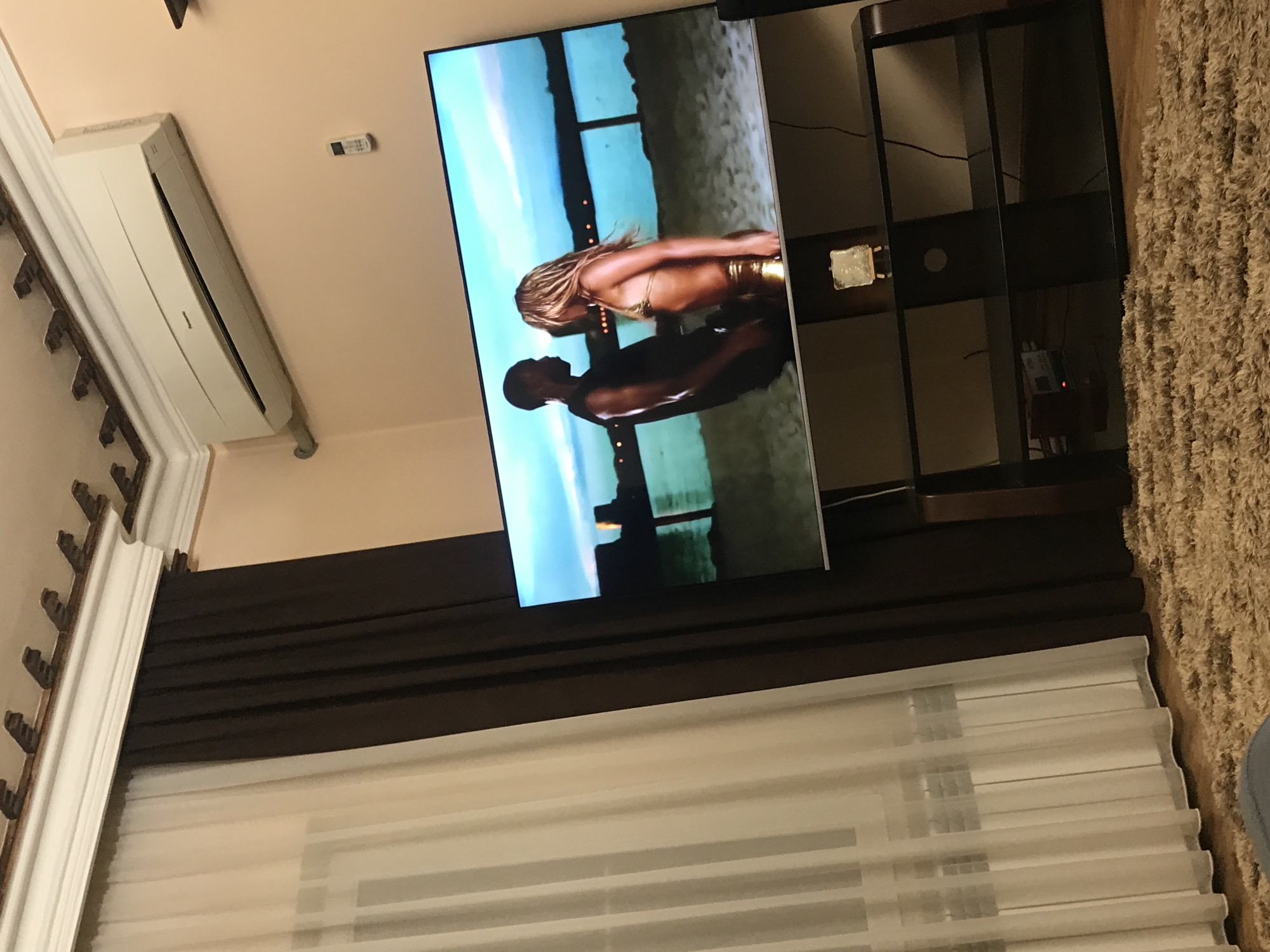 The image shows an interior view of a room. A flat-screen television is displaying a scene of a couple on a beach. The television is placed on a dark-colored TV stand. To the left of the TV is an air conditioning unit mounted on the wall, and below the TV, there are window blinds. A section of a shaggy rug is visible on the right. The city and country, Yerevan, Armenia, is not discernible from the image.