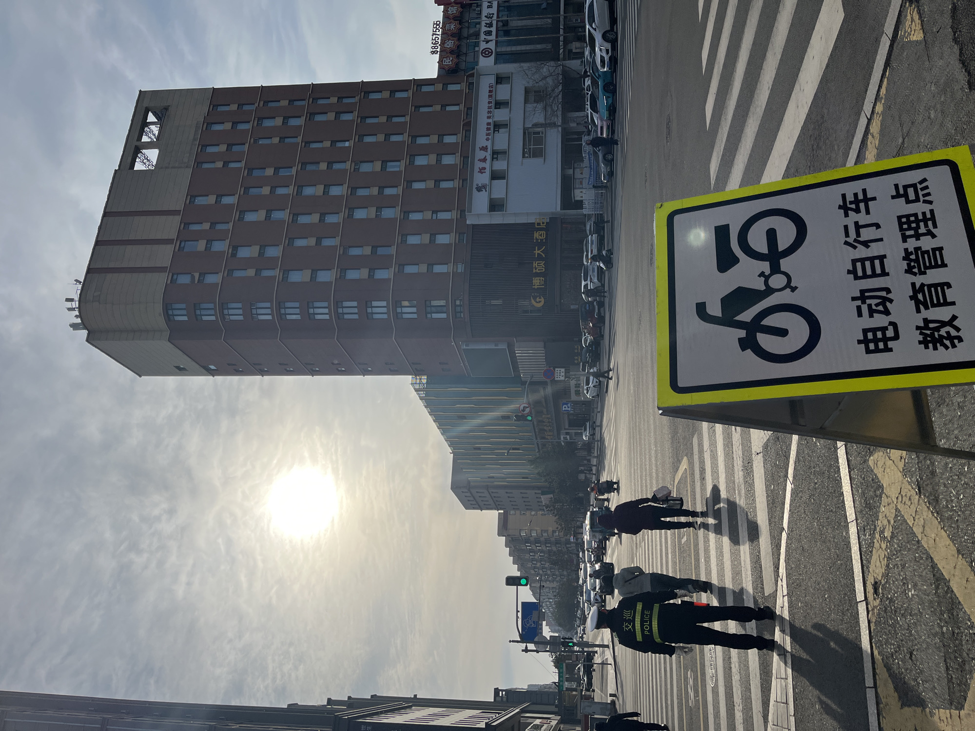 The image captures a street scene in Changchun City, China. A tall building with a brown facade stands prominently on the left. A signpost with a bicycle symbol and Chinese text, translating to "Electric Bicycle Education Management Point," is on the right. Pedestrians, including a police officer wearing a black uniform, cross a zebra crossing. Buildings line the street in the background. The sun shines brightly in the sky. Signs with Chinese characters are visible on buildings. Cars are parked along the street. The sky is overcast.