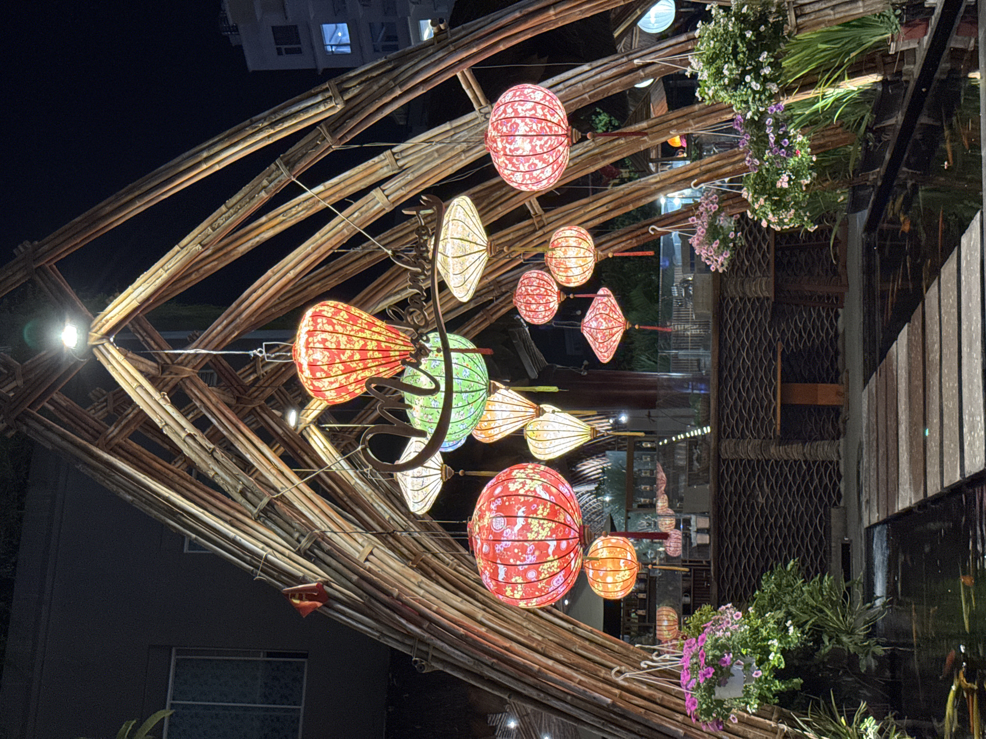 The image depicts a nighttime scene at an outdoor restaurant or cafe in Nha Trang, Vietnam, illuminated by numerous hanging lanterns of various shapes and colors, predominantly red, green, and white. A large, intricately designed bamboo structure forms an archway over a walkway, adorned with these lanterns. The lanterns have floral patterns and intricate details.
Below the bamboo structure, a dark body of water, possibly a pond or canal, is visible with koi fish swimming in it. A wooden boardwalk with planks crosses the water. On either side of the boardwalk and the pond, potted plants with pink and white flowers and green foliage are arranged.
In the background, a low structure with patterned walls and a wooden table is visible, suggesting seating or a bar area. The overall ambiance is festive and decorative, typical of many establishments in Vietnam, especially those catering to tourists. The surrounding buildings in the distance are dimly lit, indicating it's a developed urban area. There are no people clearly visible in the frame.