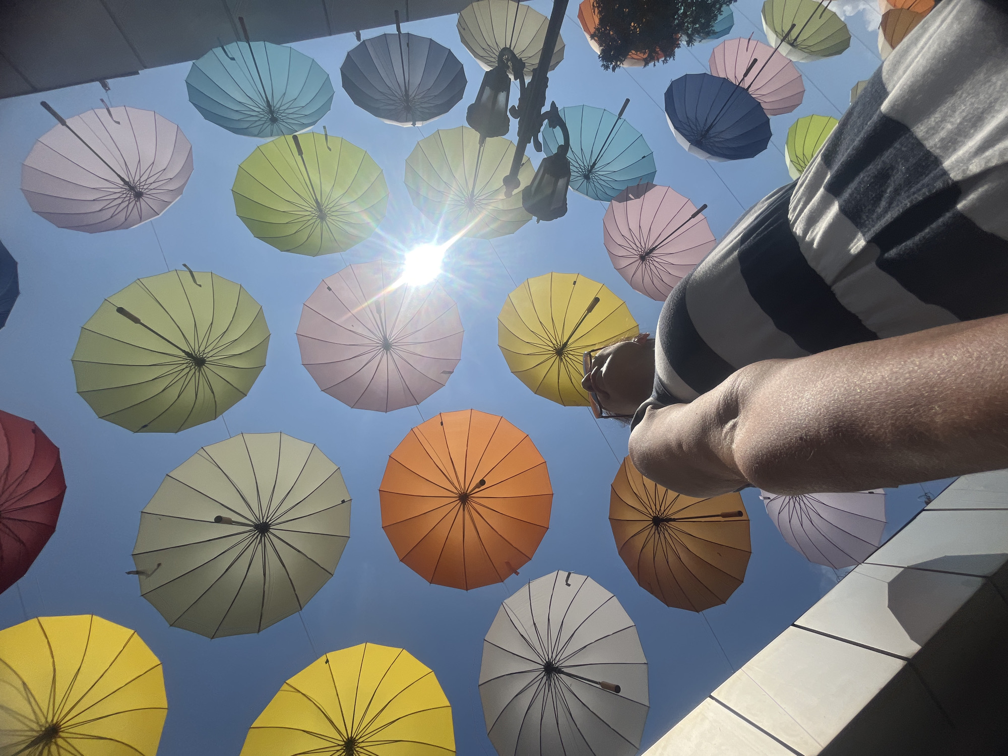 The image, taken outdoors on a sunny day, showcases a vibrant display of colorful umbrellas suspended in the air against a clear blue sky. The perspective is from below, looking upwards, emphasizing the multitude of umbrellas in varying hues of yellow, orange, pink, purple, green, and blue. Some umbrellas are closer to the viewer, appearing larger and more detailed, while others are further away and smaller. The sun is prominently visible, creating lens flare and adding to the bright atmosphere.
In the foreground, a person's arm and shoulder are visible, suggesting they are observing the umbrella installation. The person is wearing a horizontally striped dress with dark blue and white bands. The background features hints of a city environment, with what appear to be building structures on the right side and a modern architectural element with shadows on the left. The presence of streetlights suggests an urban setting.
This scene is characteristic of the "Umbrella Sky Project" installations, often found in pedestrian zones and public spaces to add visual appeal and shade. While the image itself doesn't explicitly state the location, similar installations are known to exist in various cities worldwide. This particular arrangement of umbrellas is reminiscent of displays seen in pedestrian areas of resort towns or city centers. Without further information or context, it is not possible to definitively confirm the location as Sochi, Russia.