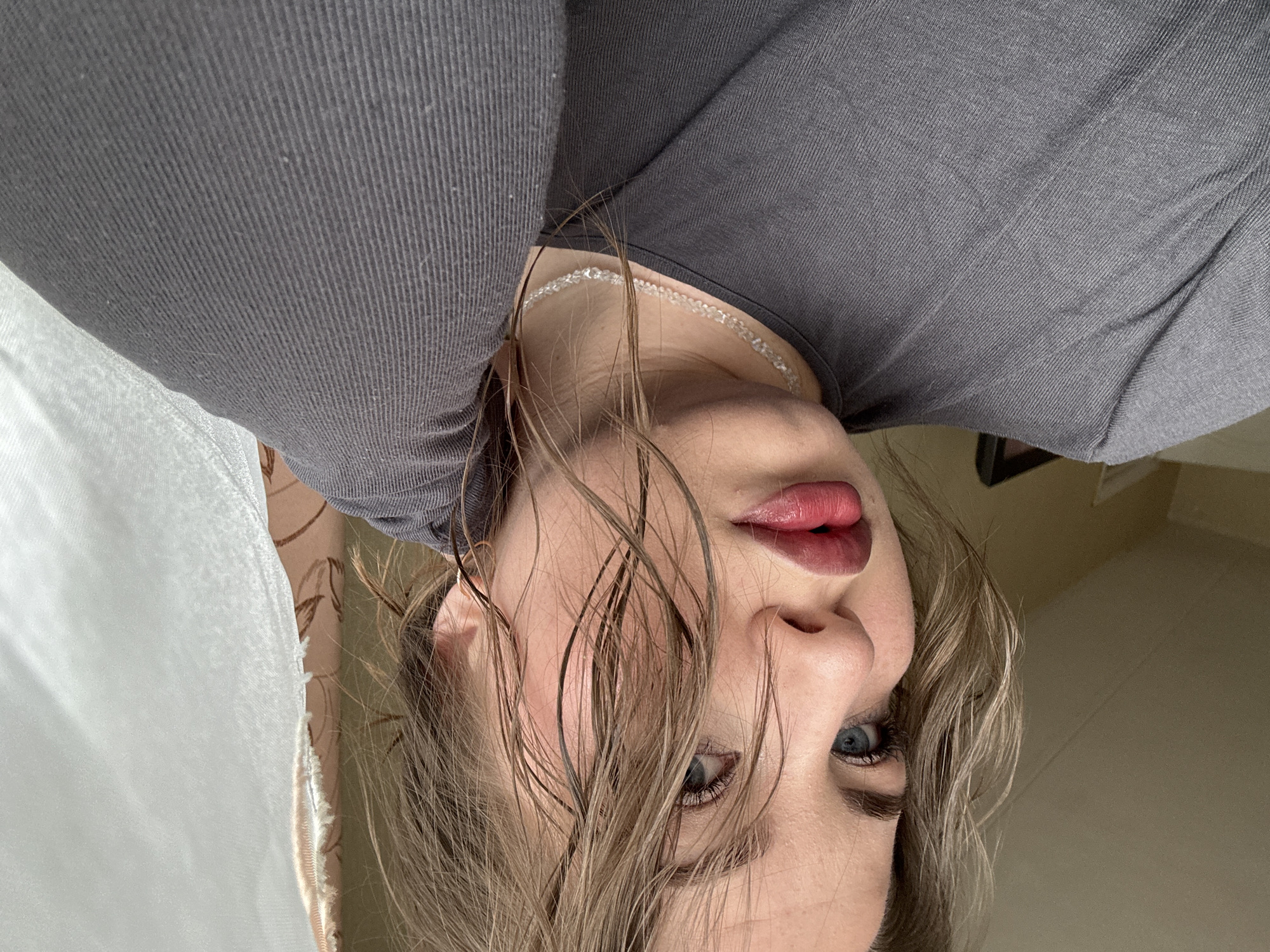 A person with wet, blonde hair is seen in a close-up portrait. They are wearing a gray top and a delicate, possibly beaded, necklace. Their lips are coated with a dark red lipstick, and they have blue eyes. A sheer, white curtain is visible in the background. The exact location in the city of Chaikovsky, Russia cannot be determined from the image alone.