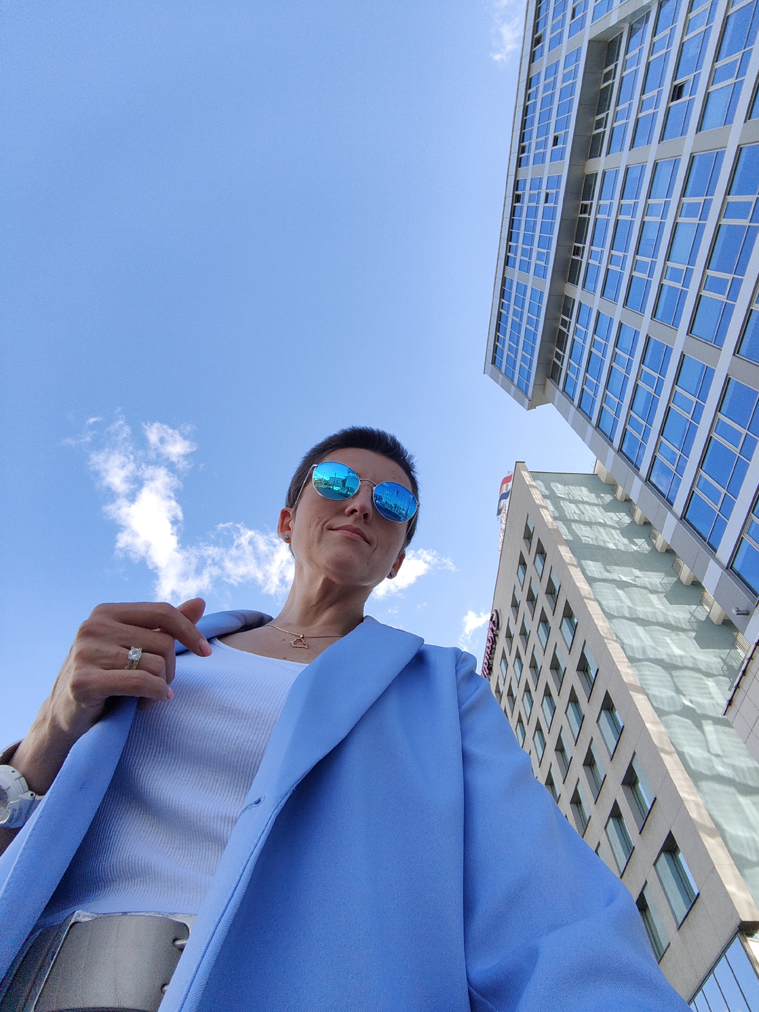 The image shows a low-angle, full-body shot of a woman standing outdoors in Minsk, Belarus, against a clear blue sky with scattered clouds. She is wearing a light blue blazer over a white ribbed tank top and a silver belt. She has short dark hair and is wearing mirrored blue sunglasses, a delicate necklace with a pendant, a ring on her left hand, and a watch on her left wrist. Her left arm is bent, and her index finger is pointing towards the camera. In the background, two modern high-rise buildings with many windows are visible, angled upwards towards the sky. The building on the right is taller and has a facade primarily composed of blue glass. The building to its left has a lighter-colored facade with a grid of windows. The overall scene conveys a sense of urban sophistication.