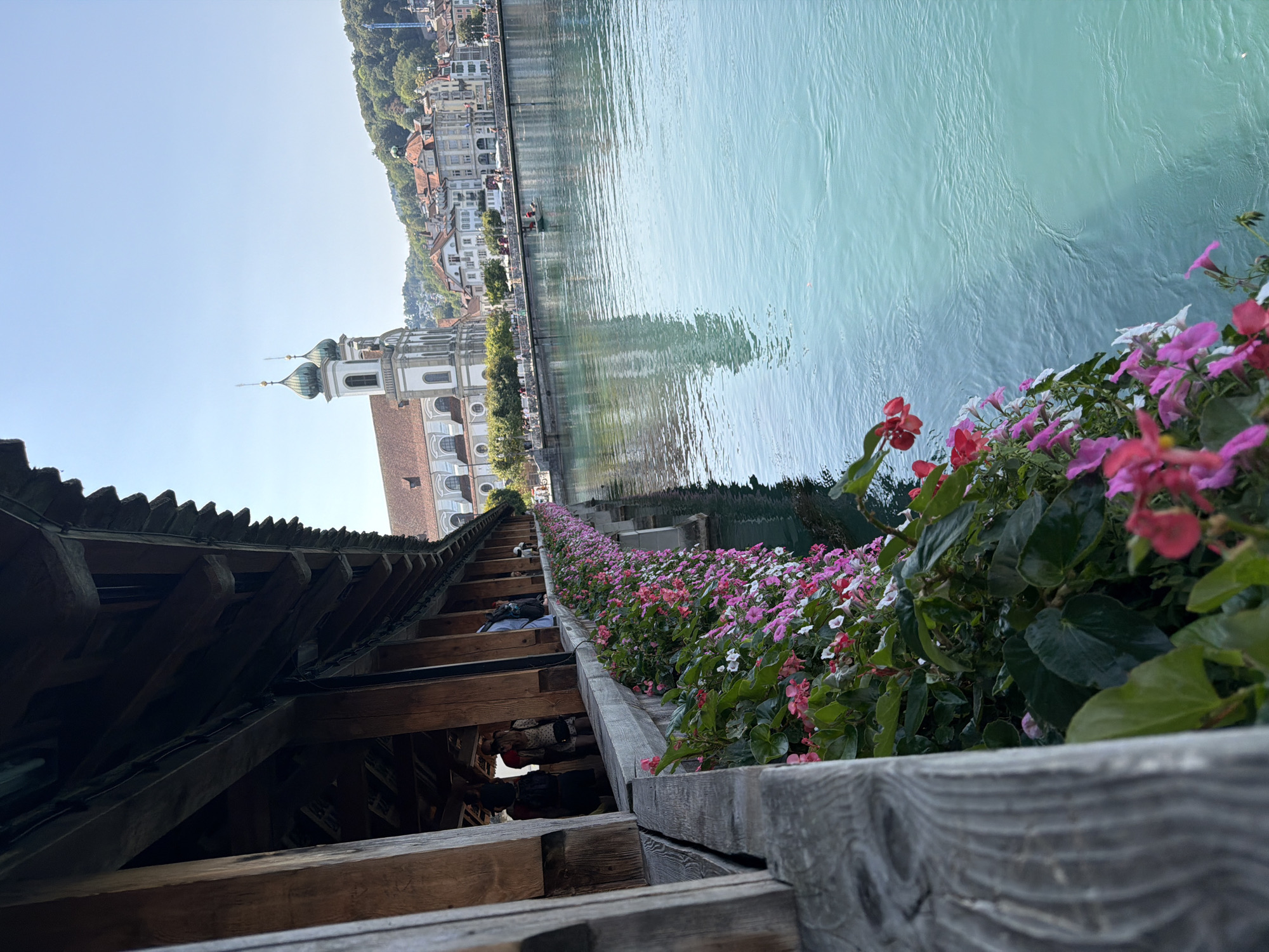 The image depicts a picturesque view of a wooden bridge adorned with vibrant pink and white flowers cascading over its edge. The bridge spans across a body of tranquil, turquoise water, reflecting the clear blue sky. In the background, a charming city skyline features a prominent church with two green-domed towers, surrounded by buildings with red-tiled roofs. Lush green hills rise behind the city. A small boat with two occupants is visible on the water, adding a touch of life to the scene. Several people are casually strolling along the bridge and the waterfront, suggesting a pleasant day out in a European city. The architectural style and scenic beauty strongly suggest a European location, likely a city in Switzerland, given the iconic covered bridge and the clear, vibrant water.