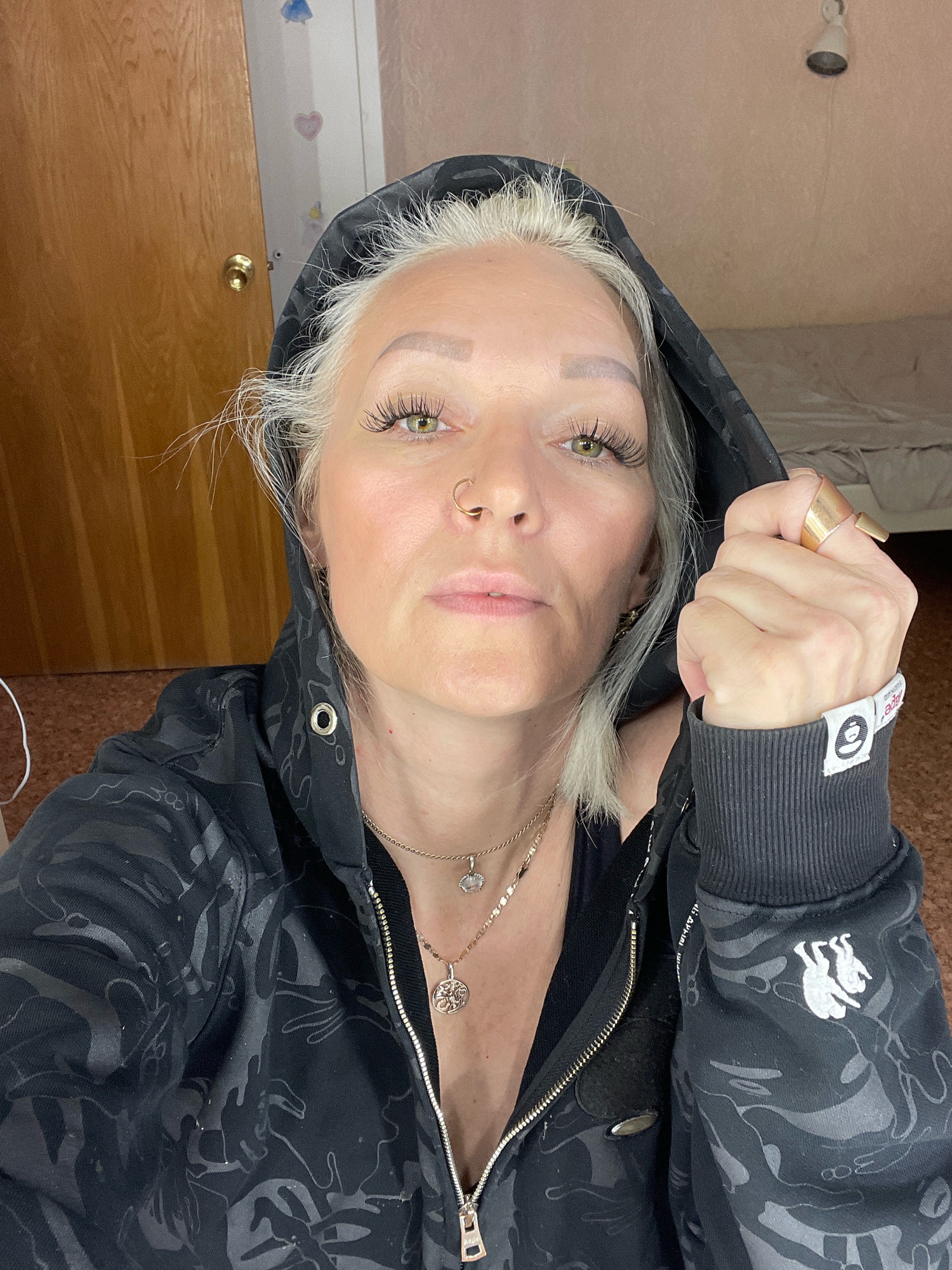 A blonde woman, wearing a black hooded jacket with a pattern, and two necklaces, is sitting on the floor in front of a wooden door and a wall with two cartoonish hearts. She is wearing false eyelashes and a nose ring. She is holding the hood of her jacket with her right hand. Her left hand, which is wearing two rings, is also visible. The woman's head is tilted slightly downwards. The background appears to be a bedroom with a bed covered with a grey blanket and a light fixture on the ceiling. The scene does not provide any indicators to suggest its location in Saint Petersburg, Russia.
