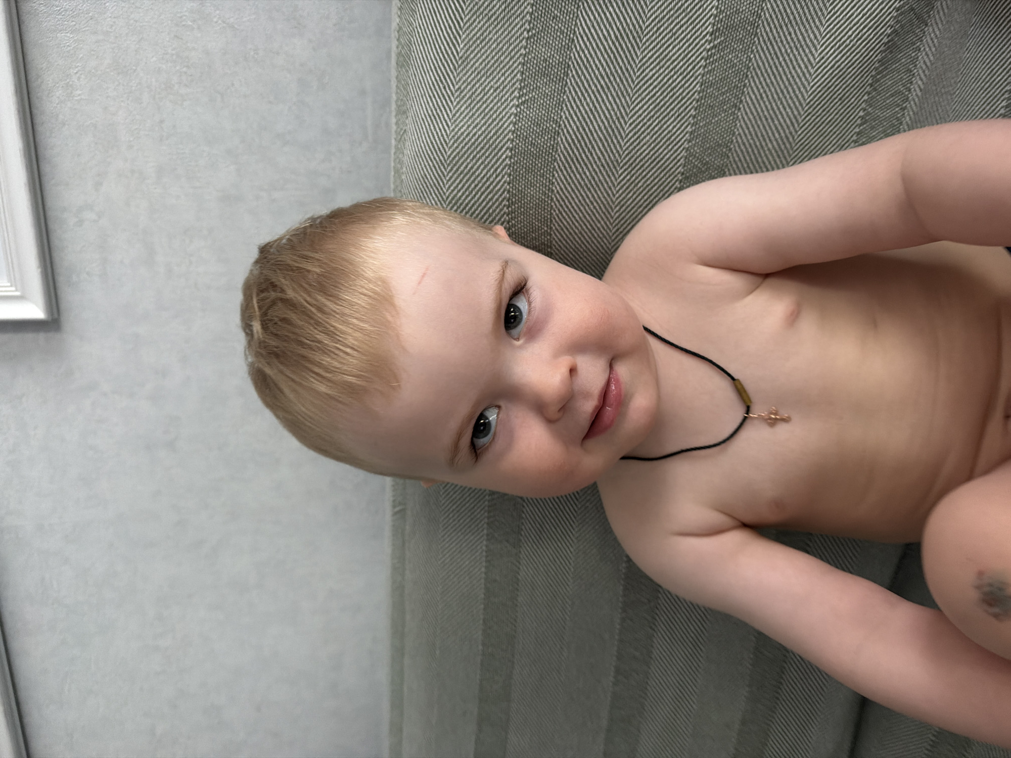 A fair-skinned, blond-haired toddler with blue eyes is lying on a green and gray herringbone patterned cushion, possibly a sofa or couch, indoors. The toddler is wearing a black necklace with a small, gold-colored pendant, and the chest is exposed. A pale, textured wall with a white framed object is visible in the background. The photo appears to be taken inside a home, possibly in a residential area. There is no information or visual evidence to indicate that the image was taken in Votkinsk, Russia.