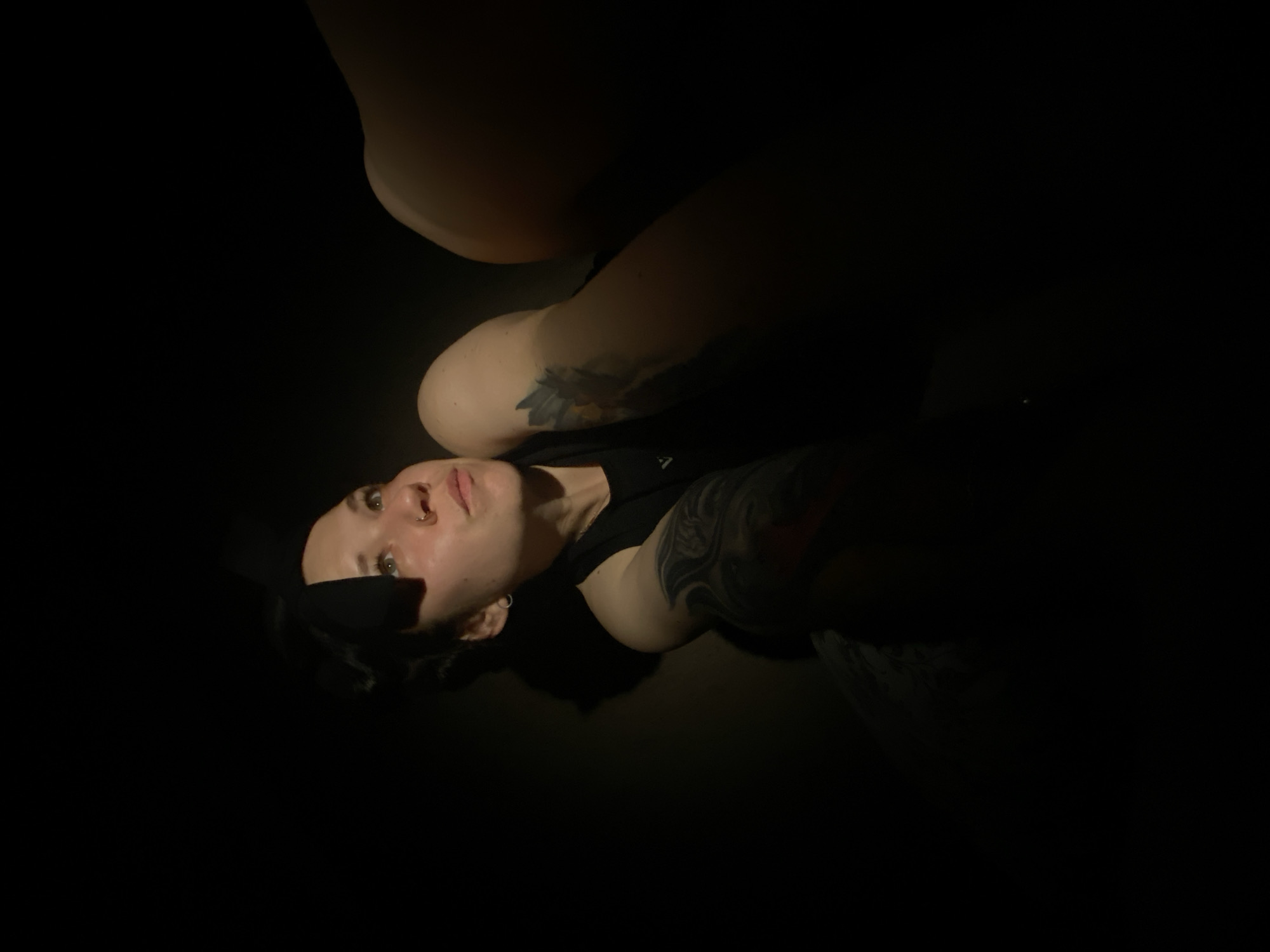A person is lying down against a dark background, illuminated by a single light source from the lower left. The person has short dark hair, fair skin, and facial piercings. The person is wearing a dark t-shirt. There are visible tattoos on the arm closest to the viewer. The background is entirely black and obscured. The location is unknown.