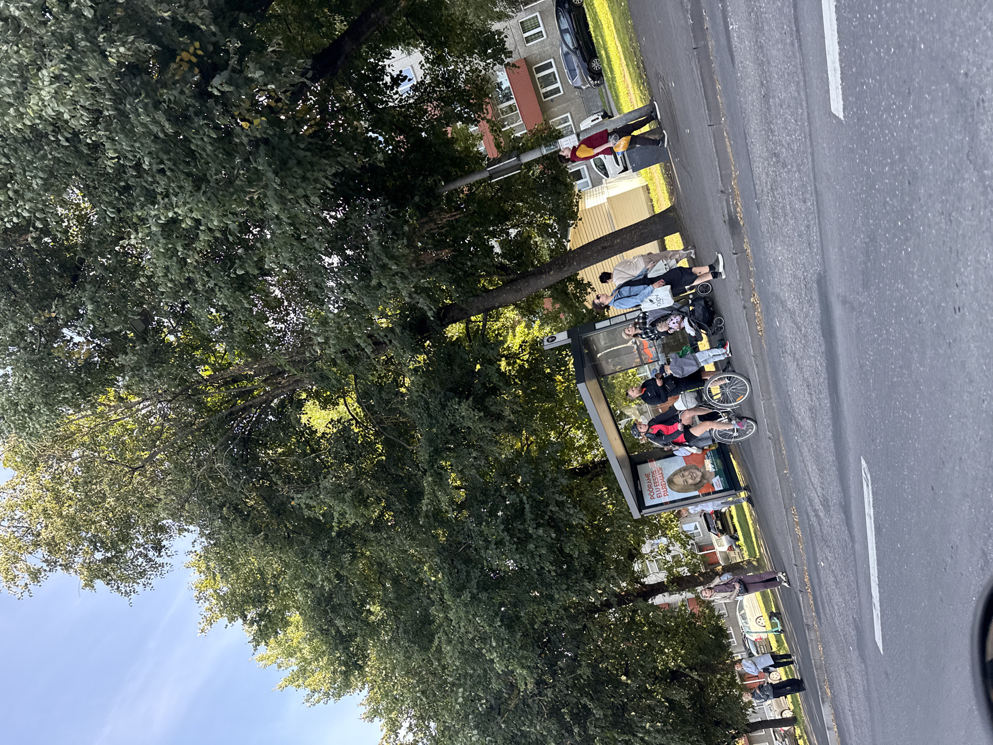 The image depicts a street scene in Tallinn, Estonia, near a bus stop on a sunny day. Several individuals are present at the bus stop and on the sidewalk. One woman is riding a bicycle, wearing a helmet and a pink jacket. Another person, also in a helmet, is walking towards the bus stop. A woman with a baby in a stroller is visible, along with other pedestrians waiting or walking by. Behind the bus stop, a billboard features a portrait of a woman with text in Estonian. To the right of the bus stop, there is a multi-story building with balconies and windows. Cars are parked on the street and in front of the building, including a black sedan and a silver car. Large trees with green foliage line the street, providing shade. The road in the foreground has lane markings.