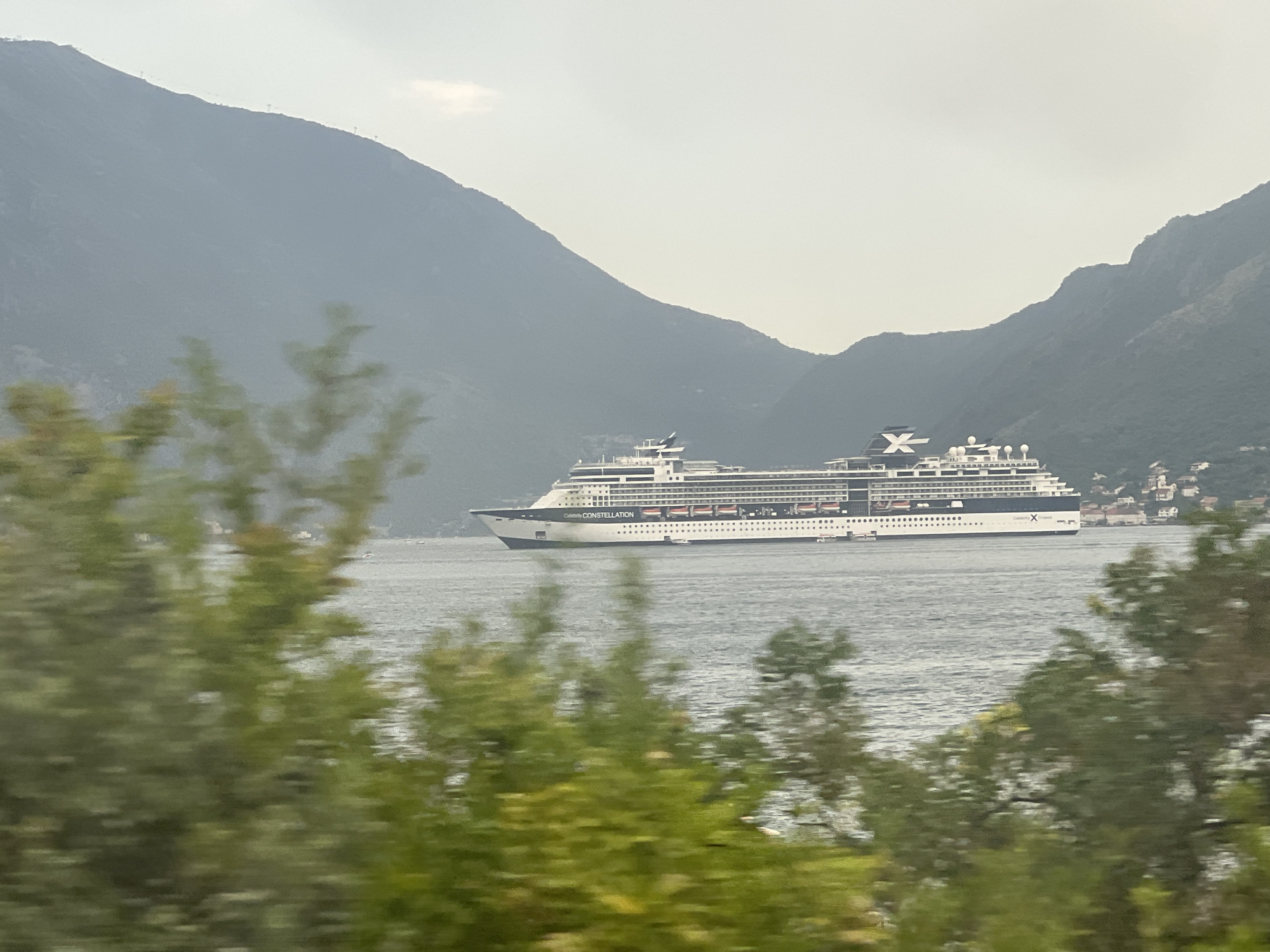 A large cruise ship, identified by the name "CELEBRITY CONSTELLATION" on its hull, sails on a calm body of water. The ship is white with black and red accents and features multiple decks with rows of windows and lifeboats. It is positioned to the right of the frame, moving left. In the background, steep, verdant mountains rise dramatically, their slopes partly obscured by a light haze. The sky above is overcast and pale. In the foreground, out-of-focus green foliage suggests a view from a moving vehicle or a point obscured by trees. The image captures a scenic view of the cruise ship navigating through a bay or fjord, with the mountainous landscape of Dražin Vrt, Montenegro, serving as a prominent backdrop.