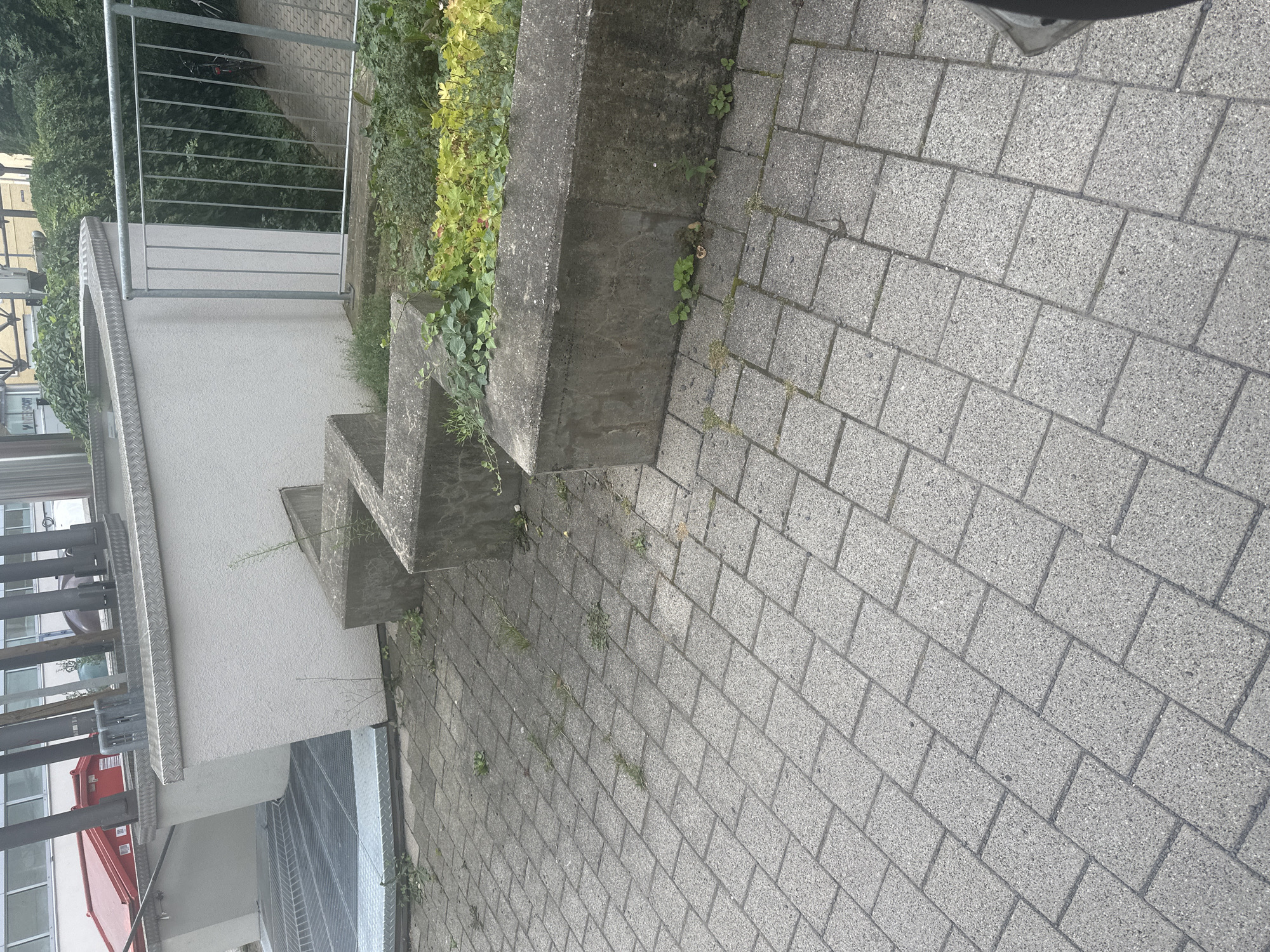 An outdoor, eye-level medium shot captures a paved walkway and a concrete planter box with foliage in the foreground. The walkway is constructed of interlocking grey rectangular pavers, some of which have weeds growing between them. A low, zig-zagging concrete planter box sits on the walkway, with sparse green plants and some yellowing foliage growing within it. The planter box has weathered concrete surfaces. In the background, a white stucco wall extends horizontally, topped with a metal railing. To the left of the wall, a metallic ramp leads downwards. To the right of the wall, a metal railing encloses an area with dense green foliage and a bicycle with a red frame parked in the background. A red dumpster is partially visible to the left of the stucco wall. The overall environment appears to be an urban outdoor setting, possibly a commercial or residential area.