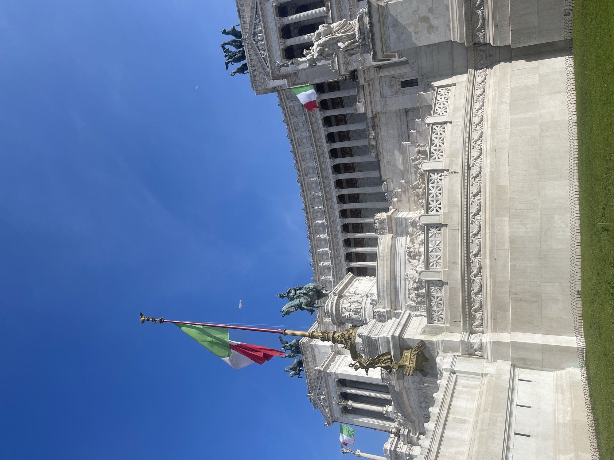 The image shows a partial view of the Altare della Patria in Rome, Italy, specifically the monument's architecture, Italian flags, and sculptures against a clear blue sky. The focus is on the intricate details of the white stone structure, including columns, reliefs, and statues. The Italian flags add a patriotic element. There is no visible presence of people. A small bird is flying in the sky.