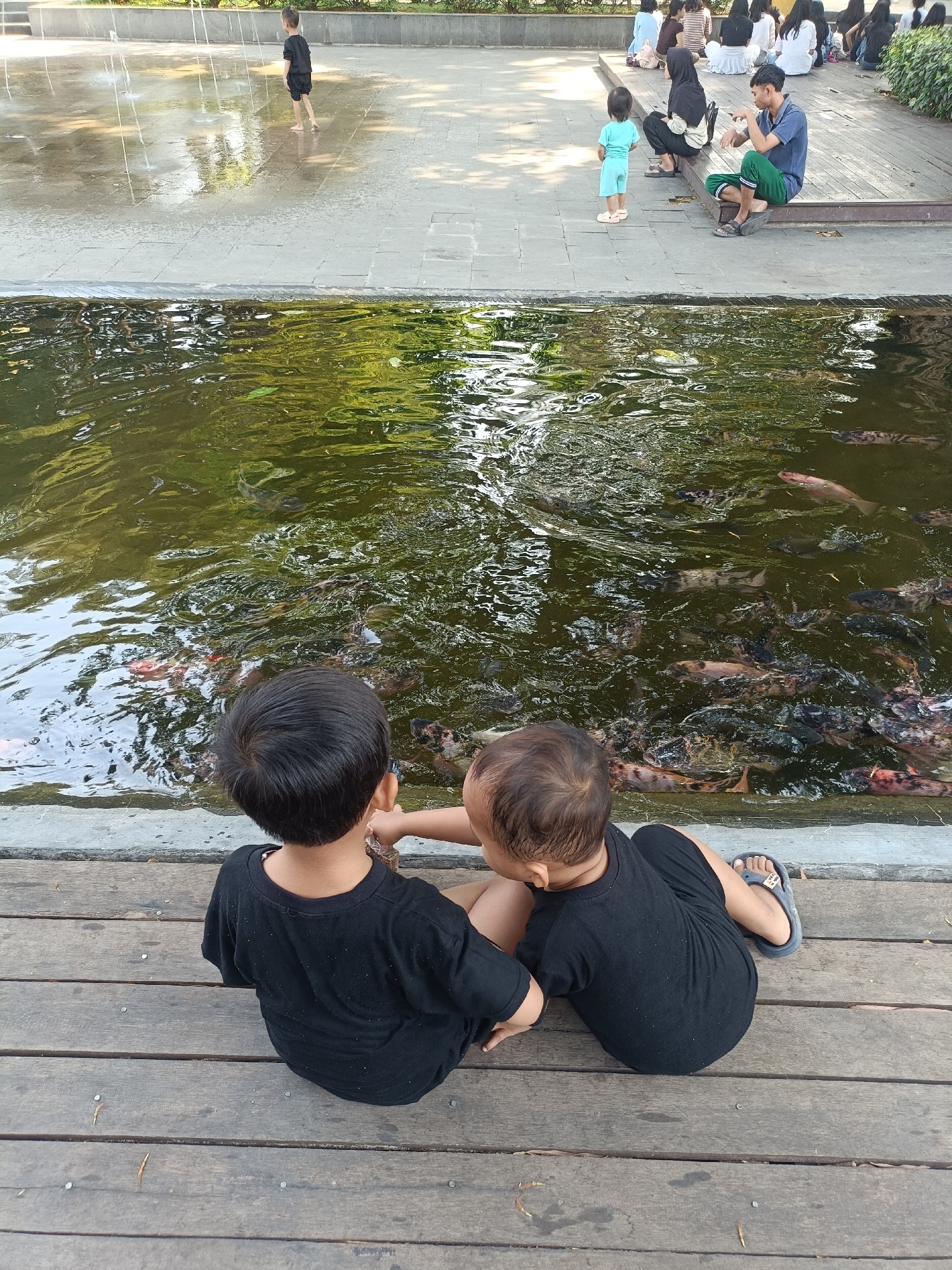 Two young children, viewed from the back, sit on a wooden deck overlooking a pond filled with many fish. Both children are wearing black t-shirts. The child on the left has dark hair, and the child on the right has lighter hair and is wearing a sandal on their right foot. The pond is murky green with visible reflections of trees and the sky. In the background, a group of people, including adults and children, are visible on a paved area near a fountain. One man in a blue shirt and green pants is sitting and talking to a small child in a light blue outfit. Other individuals are seated or standing further back. The setting appears to be a public park or recreational area with water features. The presence of the fountain, paved walkways, and abundant fish in the pond suggests this location is likely Taman Brantas in Kediri City, Indonesia, a popular spot for families and recreation.