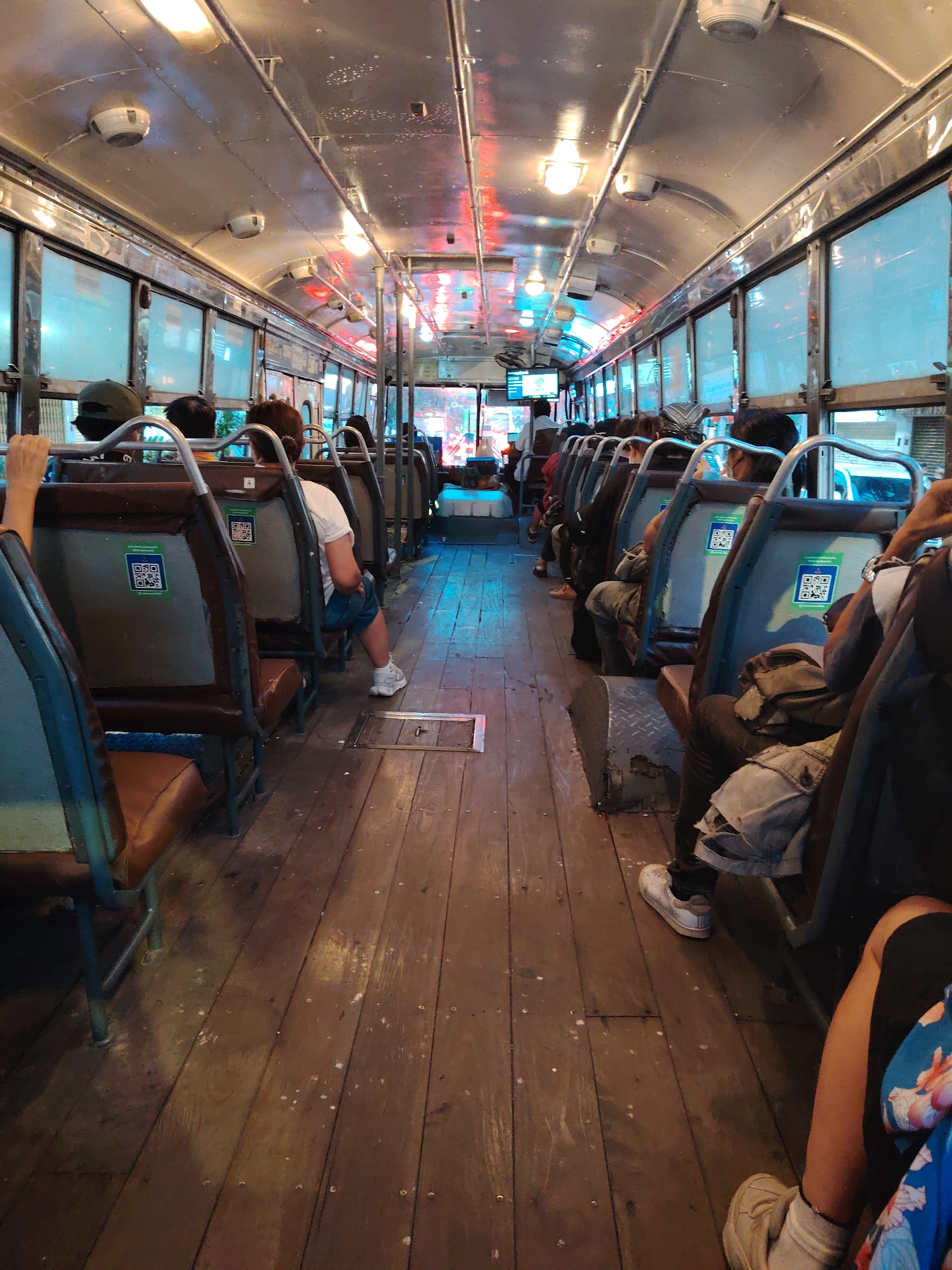 The image shows the interior of a public bus in Bangkok, Thailand, looking down the central aisle towards the front. The bus features a utilitarian design with a worn wooden plank floor and a metallic ceiling equipped with fluorescent strip lights and circular vents.
Numerous passengers are seated on both sides of the aisle, primarily viewed from behind or the side, indicating they are commuting or traveling. The seats have brown and blue upholstery with sturdy metal frames. Prominently displayed on the back of many seats are green and blue QR code stickers, accompanied by small amounts of Thai script, likely for payment or information.
Large windows line the bus, reflecting the interior lighting and offering muted glimpses of the outside urban environment, suggesting it is either evening or night, or a dimly lit day. Red and blue light reflections are noticeable on the ceiling towards the front, possibly from exterior streetlights or signage. The passengers appear to be quietly engaged in their journey, with no strong emotions discernible.