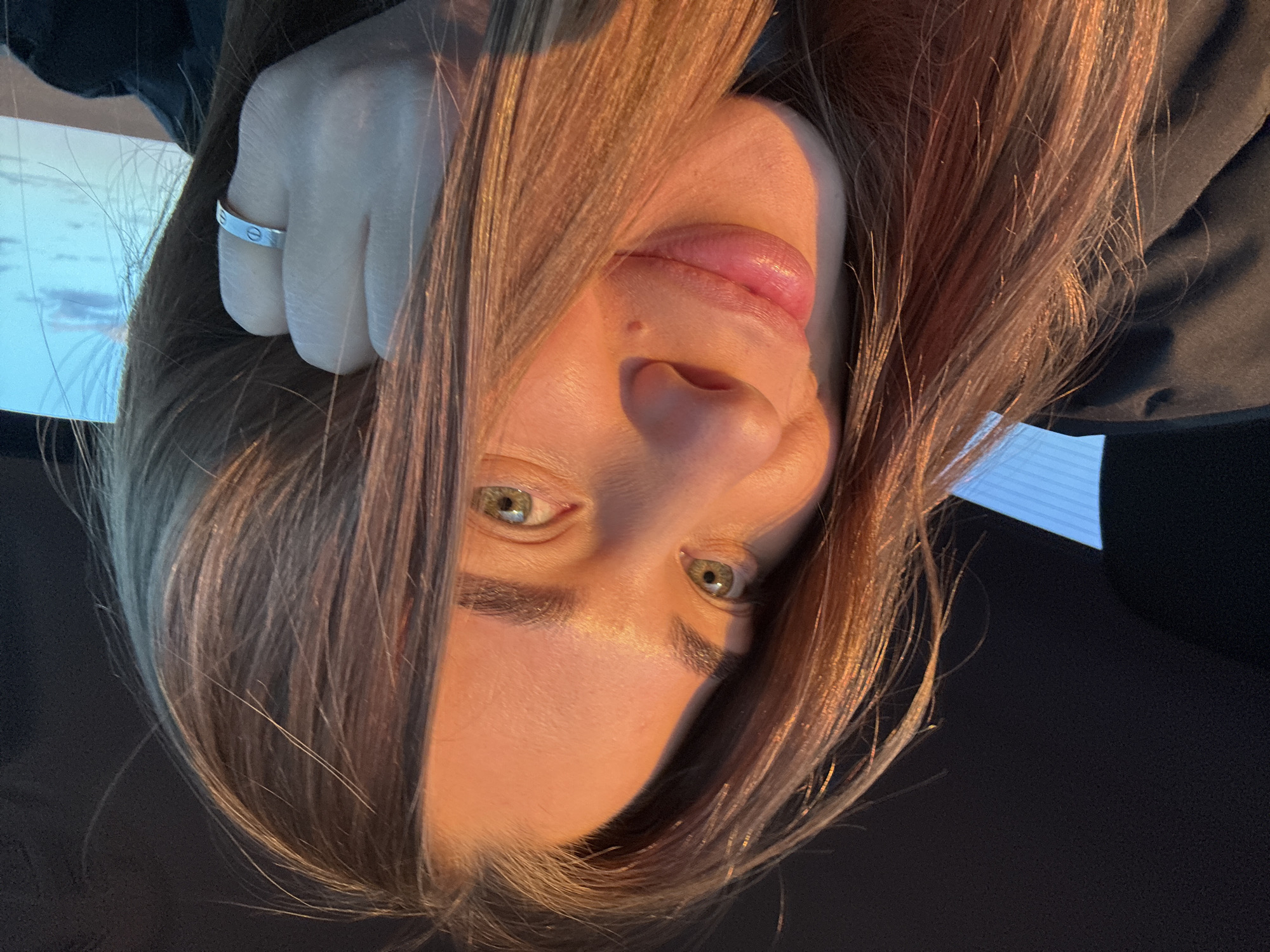 The image shows a woman's face and upper body, oriented upside down. The woman has light skin, green eyes, and brown hair. She is wearing a silver ring on her left ring finger. Her clothing appears to be black. The background is dark, with a small area of bright light visible in the upper left corner. The location given is Сыйрышбашево, Russia, but the image provides no additional information to confirm the accuracy of the given location.