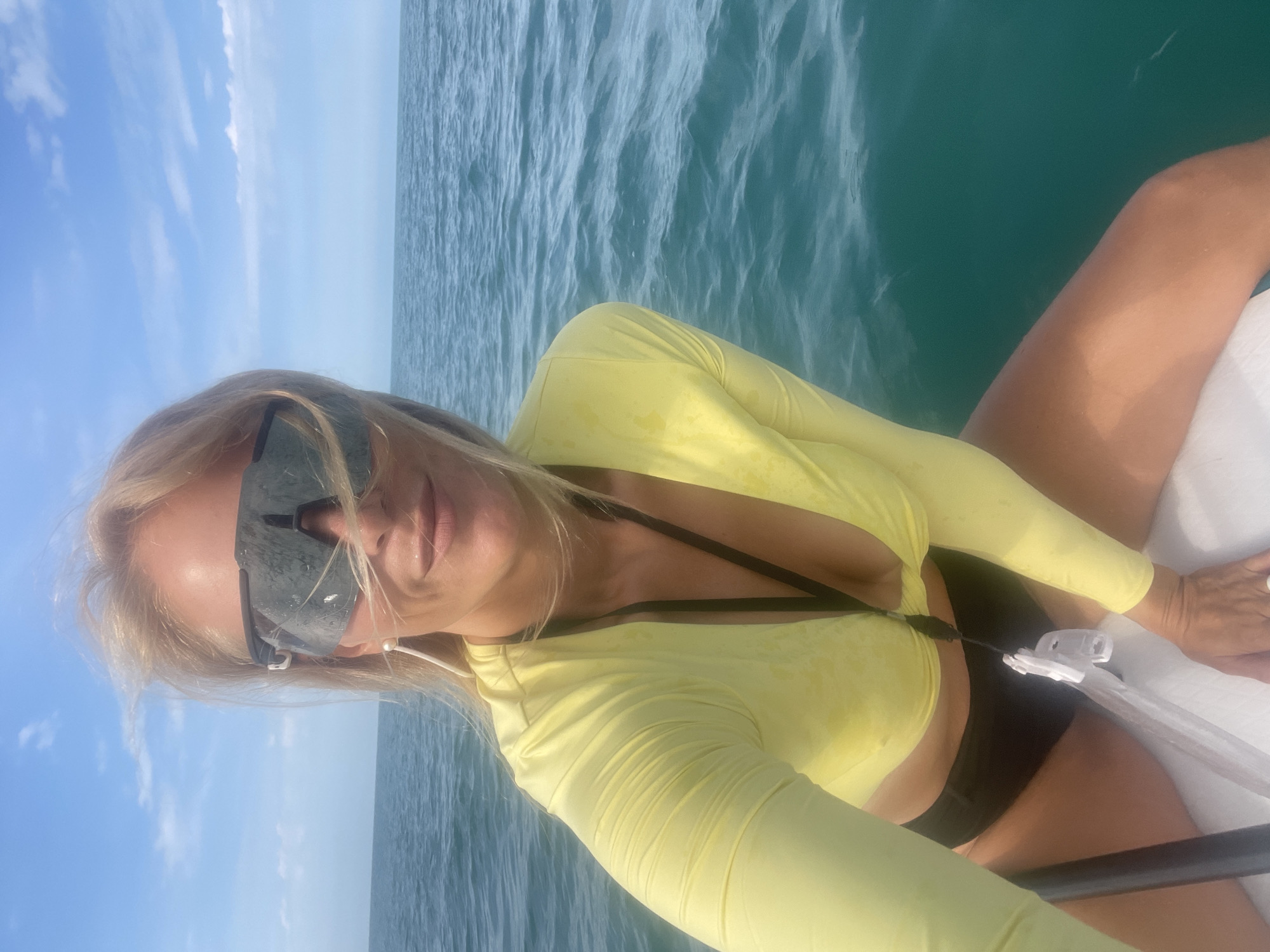The image shows a woman in sunglasses and a yellow long-sleeved crop top and black bottoms posing on what appears to be a boat. The water is blue and choppy, and the sky is bright blue with scattered clouds. The woman has blonde hair and is smiling. There is no indication of the city of Sochi, Russia in the image.
