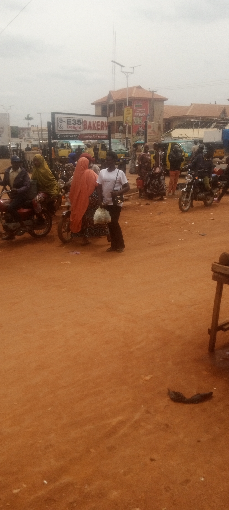 The image depicts a street scene in Sabon Tasha, Nigeria, characterized by a dusty, reddish-brown ground. In the background, a sign for "E35 Bakery" is visible along with other commercial buildings and advertisements. People are interacting near parked motorcycles. Some are wearing traditional clothing and hijabs. A woman in a white t-shirt stands holding a bag. Yellow taxis are parked behind them. The sky is overcast. A wooden structure is on the bottom right corner of the frame.