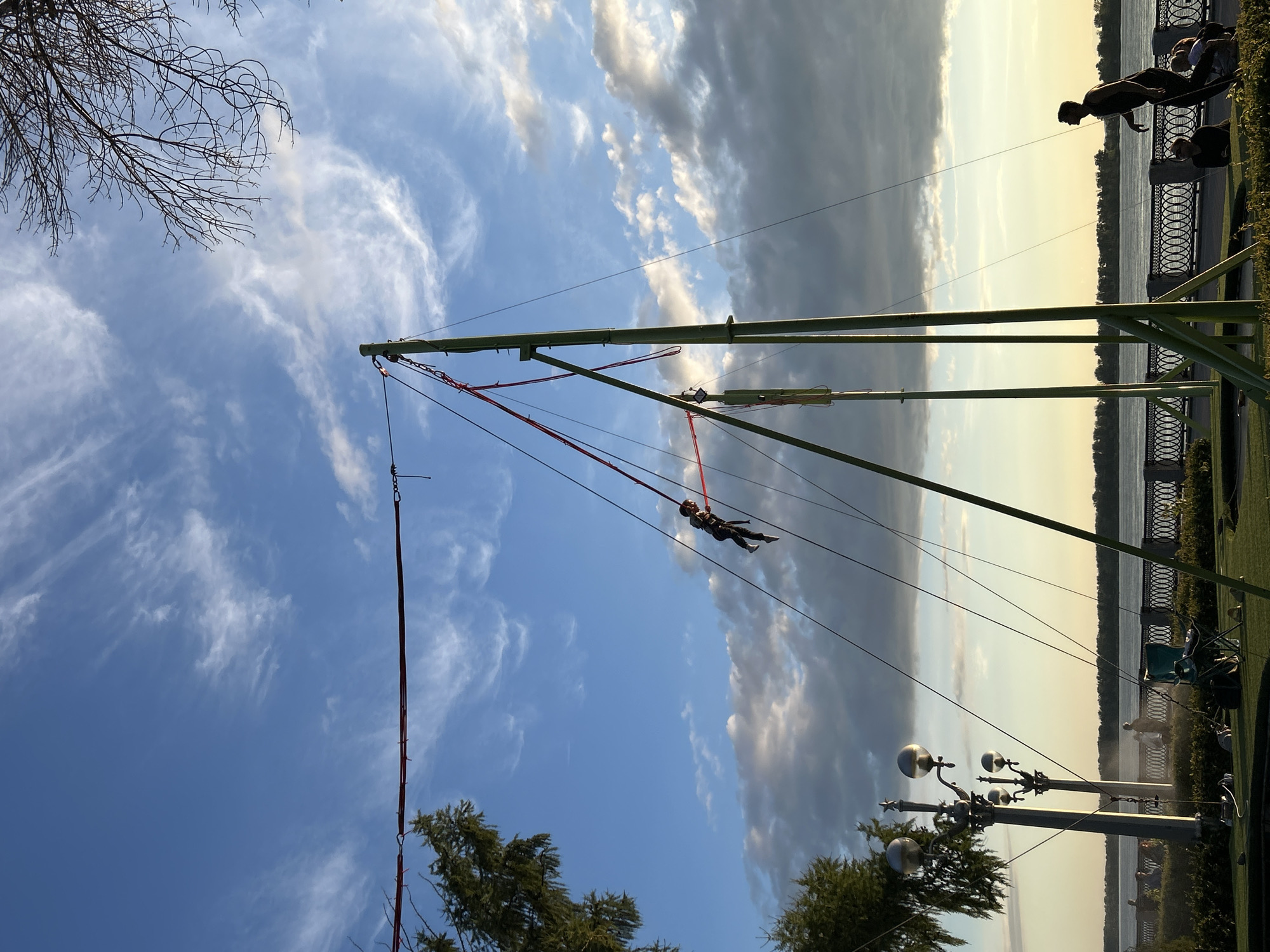 The image captures a vibrant outdoor scene against a backdrop of a dramatic sky. The primary subjects are a person suspended mid-air by bungee cords, attached to a tall green structure. This person, likely a child based on their size, is wearing dark clothing and appears to be enjoying the thrill of bungee jumping.
The setting is a park or recreational area situated beside a body of water, possibly a river or lake, with a well-maintained embankment featuring a decorative railing. In the background, silhouetted figures of other people can be seen, some walking along the embankment and others possibly waiting for their turn. A row of ornate streetlights stands along the path, adding to the scenic view. Trees, both bare branches and foliage, frame the scene, suggesting it's either early spring or late autumn.
The time of day appears to be late afternoon or early evening, as indicated by the soft, warm light and the long shadows cast by the structure and the people. The sky is a mixture of clear blue and overcast, with wispy white clouds interspersed with darker, more dramatic cloud formations, creating a visually dynamic contrast.
The overall emotion conveyed is one of exhilaration and enjoyment, with the bungee jumper clearly in the midst of an exciting activity. The presence of other people enjoying the park suggests a leisurely and pleasant atmosphere. No visible text is present in the image.