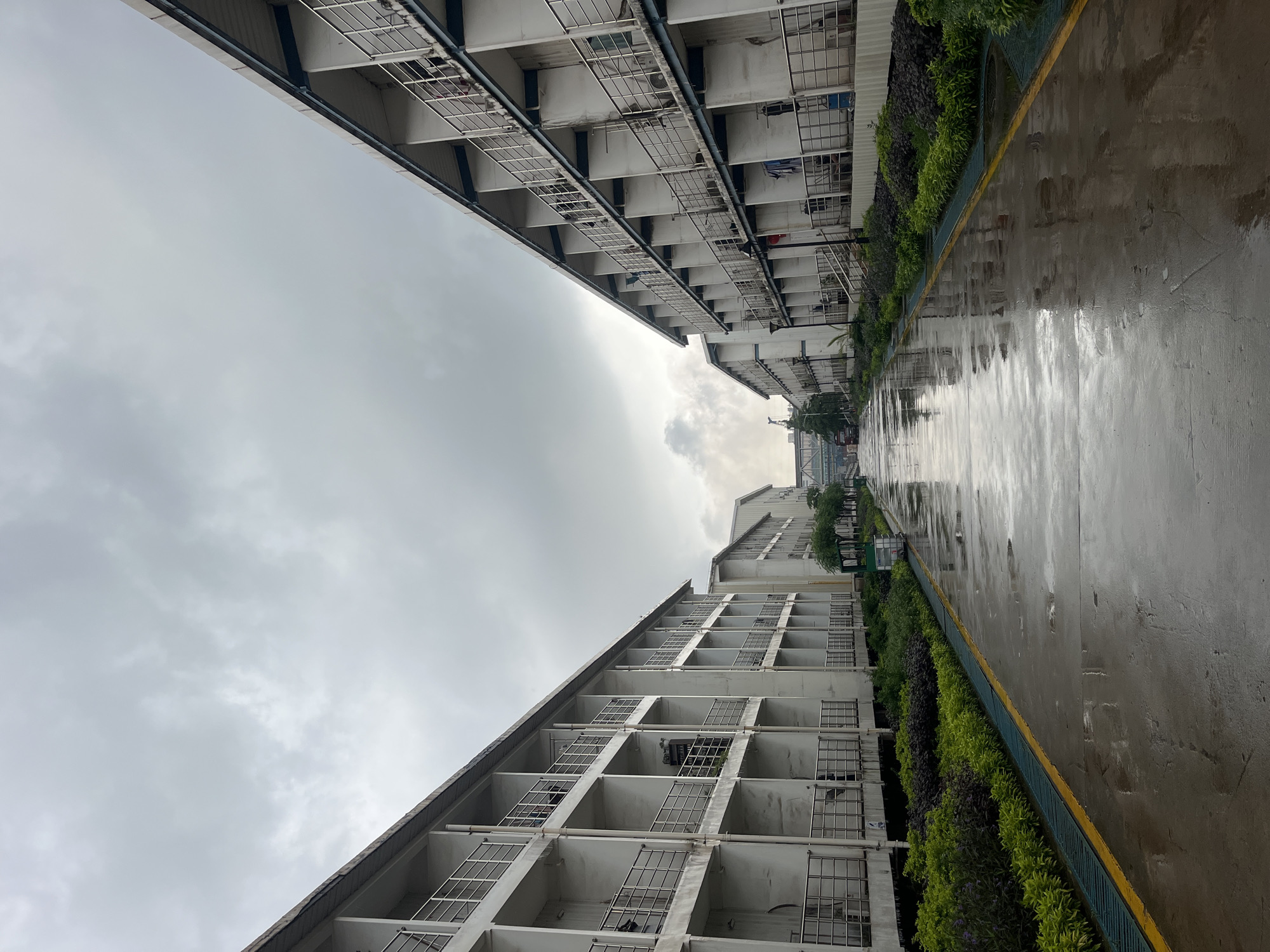 The image depicts a view of tall buildings on either side of a wet, shiny pathway or road. The buildings are multi-story and appear to have balconies or window boxes. The sky above is overcast with gray clouds. A strip of greenery or shrubbery lines the edge of the pathway next to one of the buildings. The location is possibly a street in an urban area of Unknown, Indonesia. No people are visible.