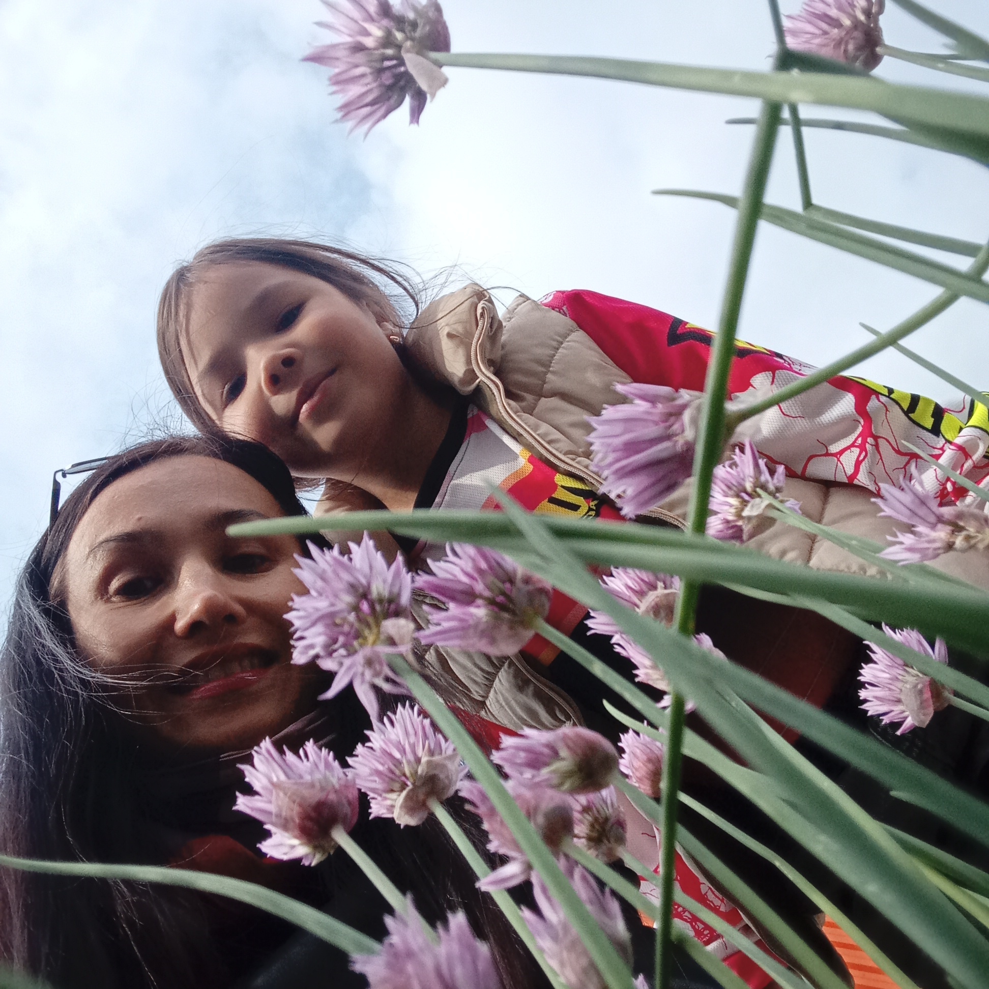 This low-angle photograph captures an adult woman and a young girl looking downwards towards the camera, framed by numerous purple chive blossoms and their vibrant green stems. The scene takes place outdoors under an overcast sky, suggesting diffused natural light during the daytime in Pervomaysky, Russia.
The woman, with dark hair and sunglasses resting on her head, displays a gentle, relaxed smile with red lipstick. The young girl, positioned slightly behind and above the woman, has light brown hair and is wearing a beige quilted vest over a colorful top patterned in red, yellow, and white. She also looks down with a calm and soft expression.
The foreground is dominated by a dense cluster of chive blossoms, some in sharp focus while others are blurred, creating a sense of depth and framing for the subjects. The upward perspective emphasizes the individuals against the pale, cloudy sky, which occupies the upper portion of the frame. The overall mood is serene and intimate, depicting a shared moment of connection. No text is visible in the image.